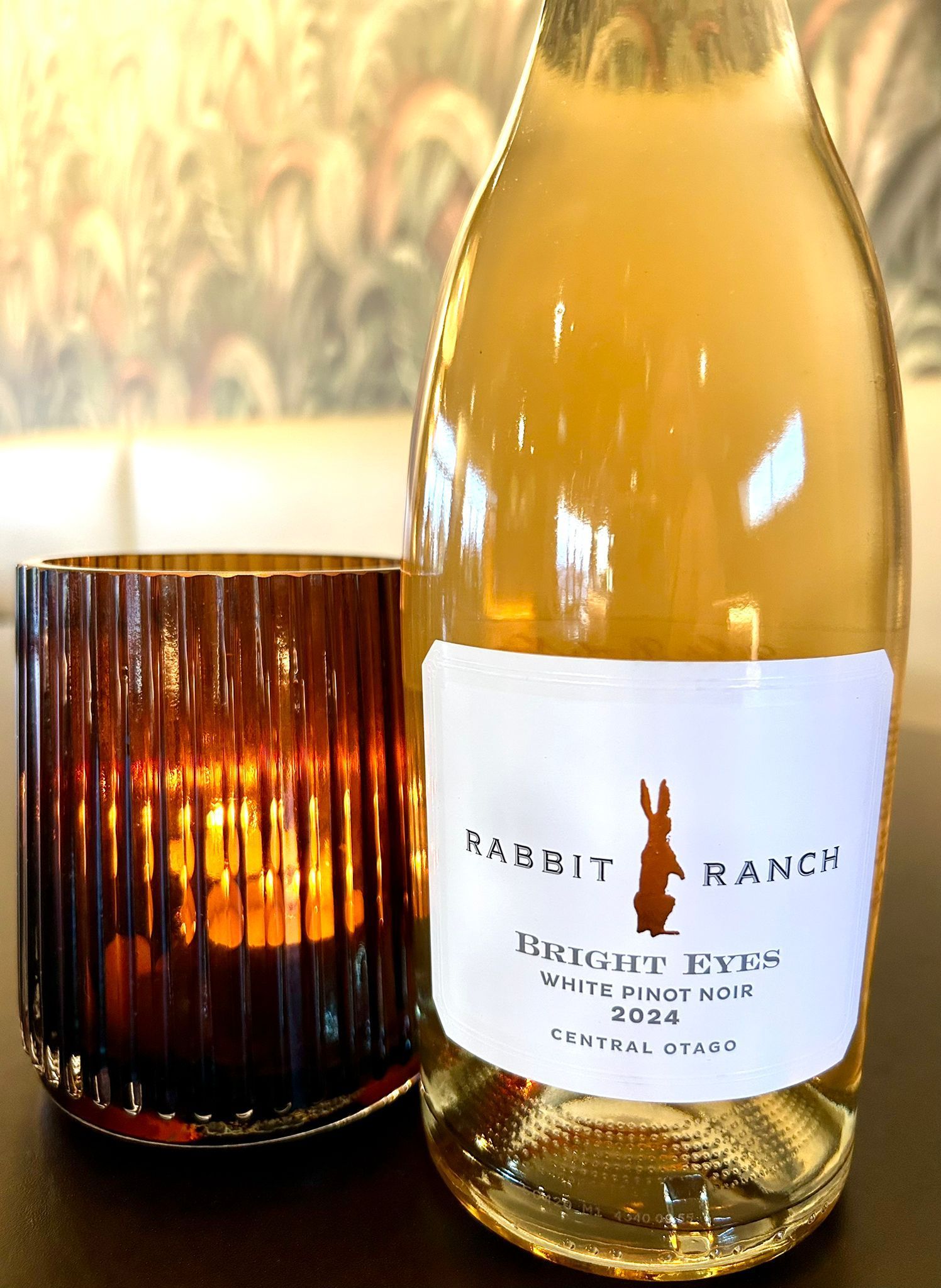 A bottle of Rabbit Ranch Bright Eyes Pinot Gris 2020 sits next to a warm, glowing ribbed glass candle holder.