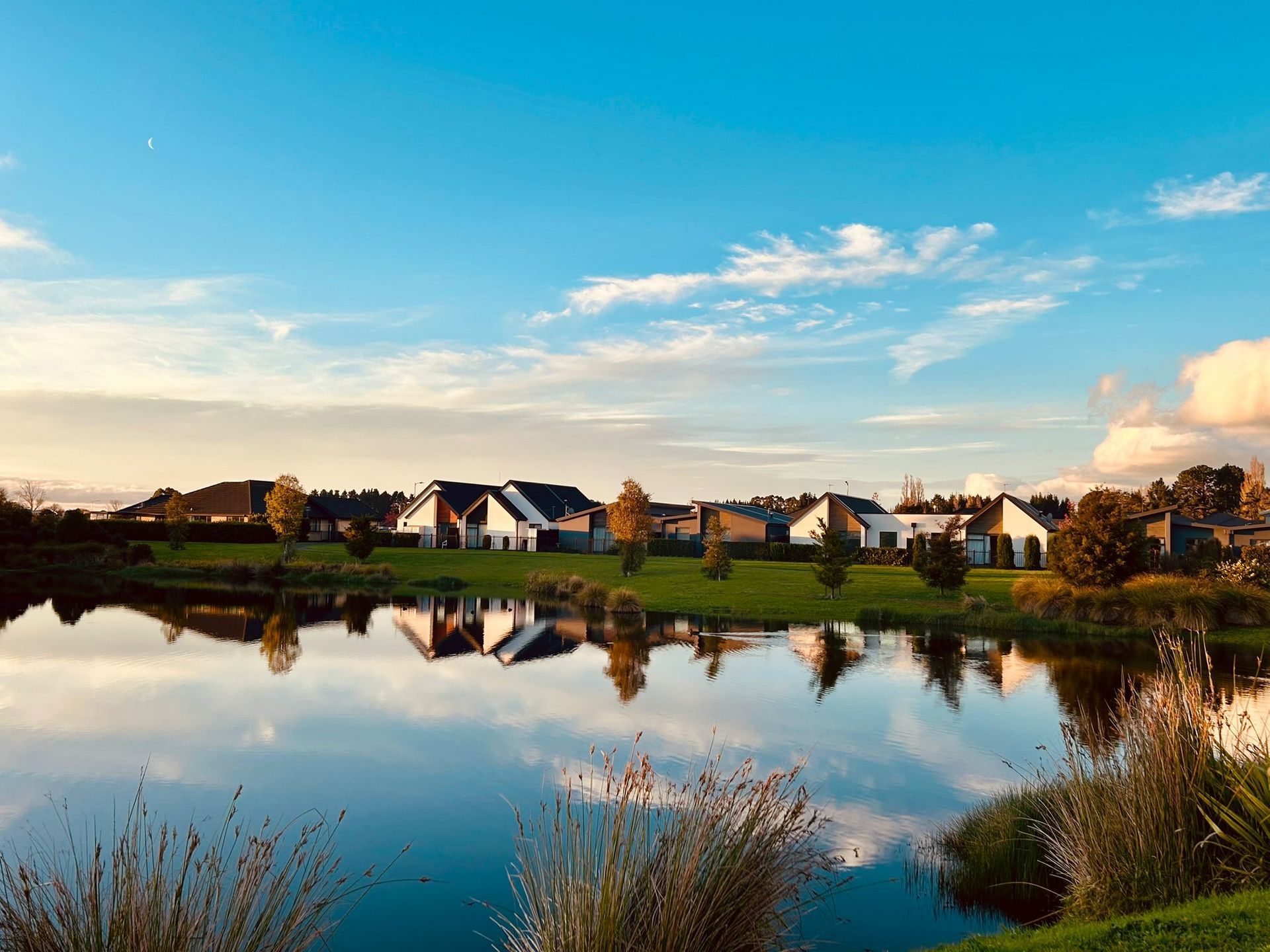 A calm pond reflects modern houses at sunset, with a vibrant blue sky and soft golden light over a grassy landscape.