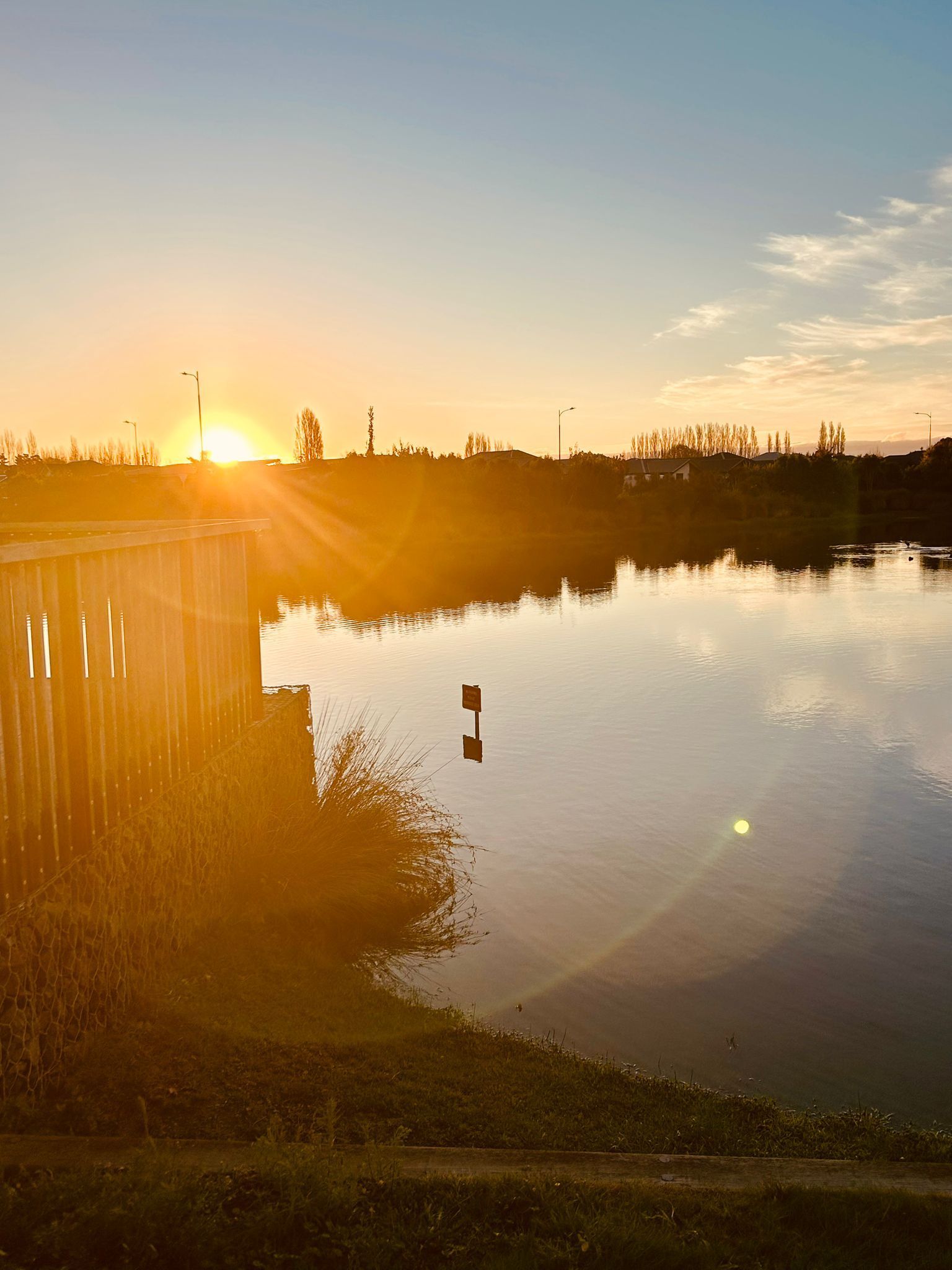 A golden sunrise glows over a calm lake, casting a lens flare and reflections near a wooden fence on the shore.