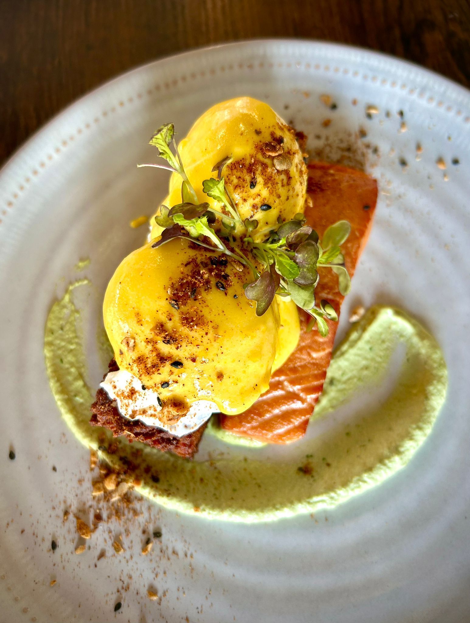 Poached eggs with hollandaise sauce on toast and salmon, garnished with microgreens and a creamy green swirl on a plate.
