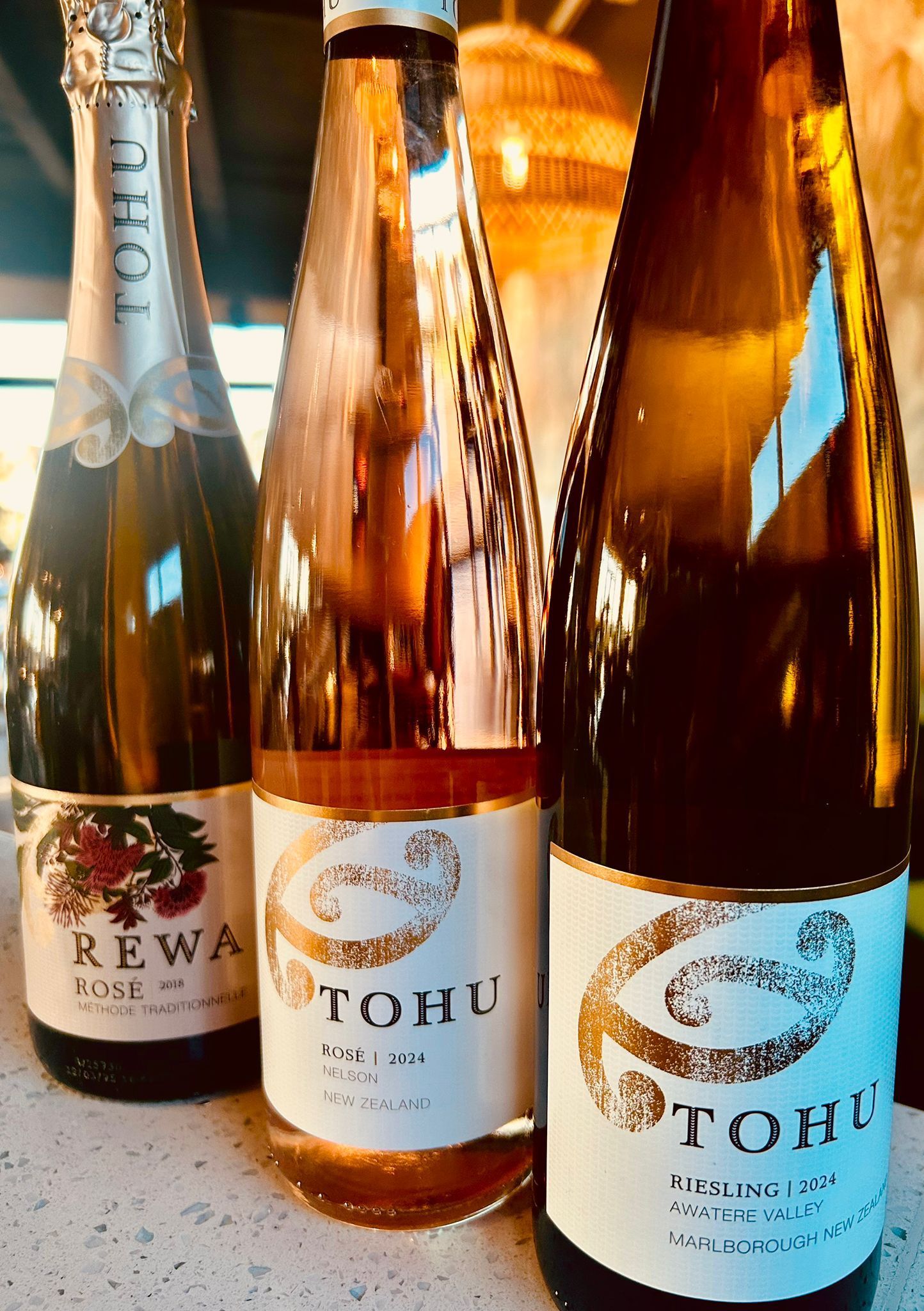 Three bottles of Tohu and Rewa wine standing on a counter in a dimly lit room.