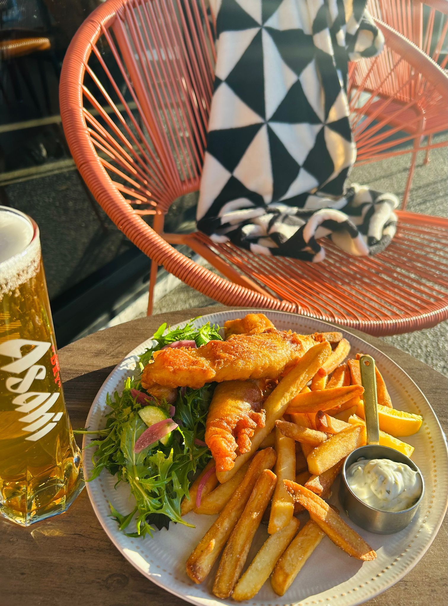 A plate of fish and chips with salad, a side of sauce, and an Asahi beer, set on a table by a bright orange chair.