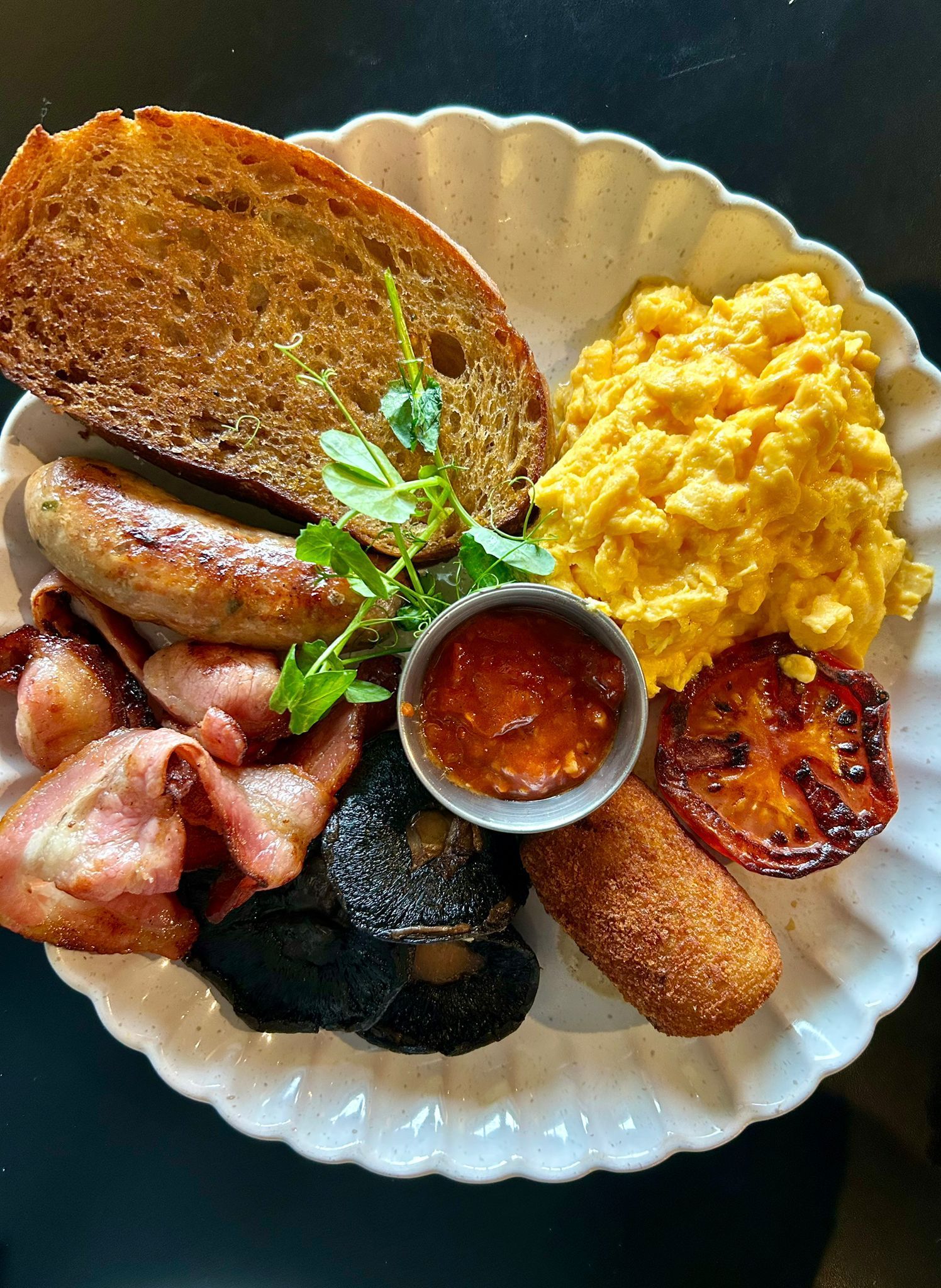 A plate containing a full breakfast with eggs, bacon, sausage, black pudding, a fried tomato, bread, and a relish cup.