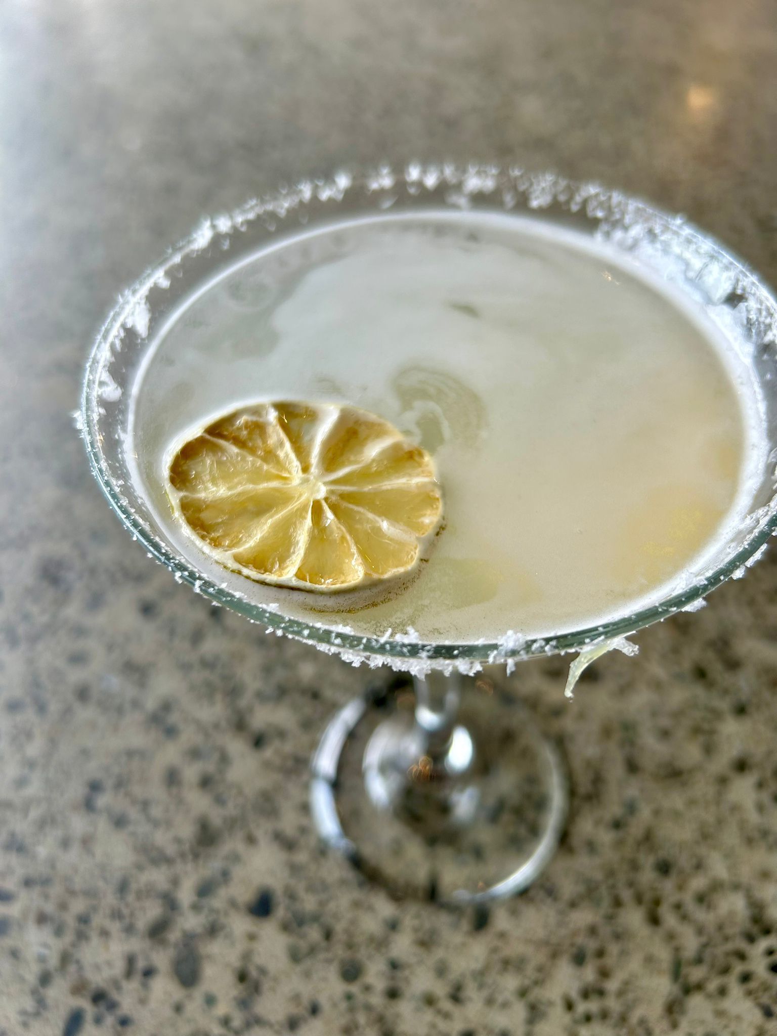 A salted-rim martini glass holds a pale cocktail garnished with a lime wheel, set against a mottled gray countertop.
