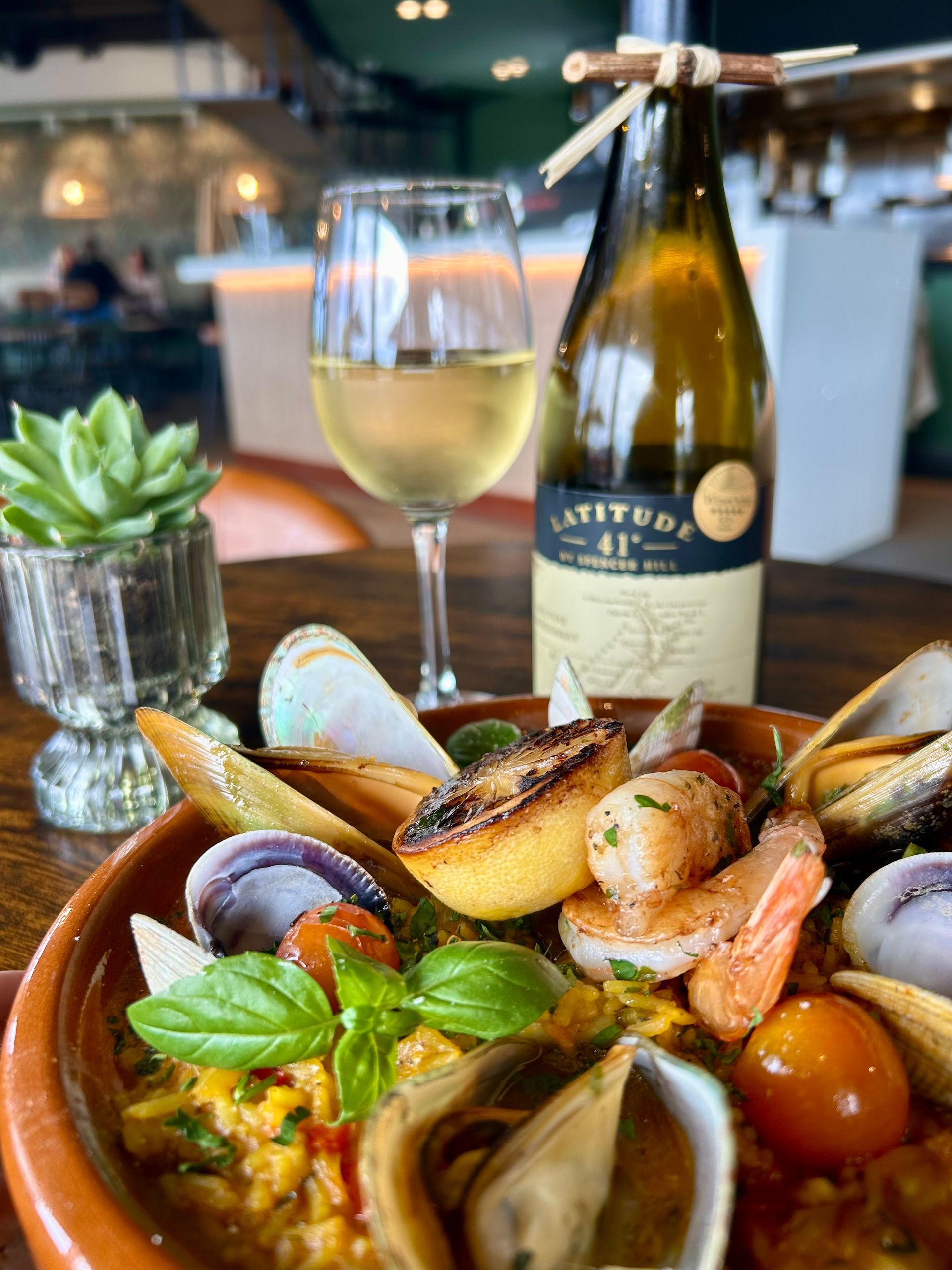 A plate of seafood paella with clams and shrimp next to a glass and bottle of white wine in a restaurant setting.
