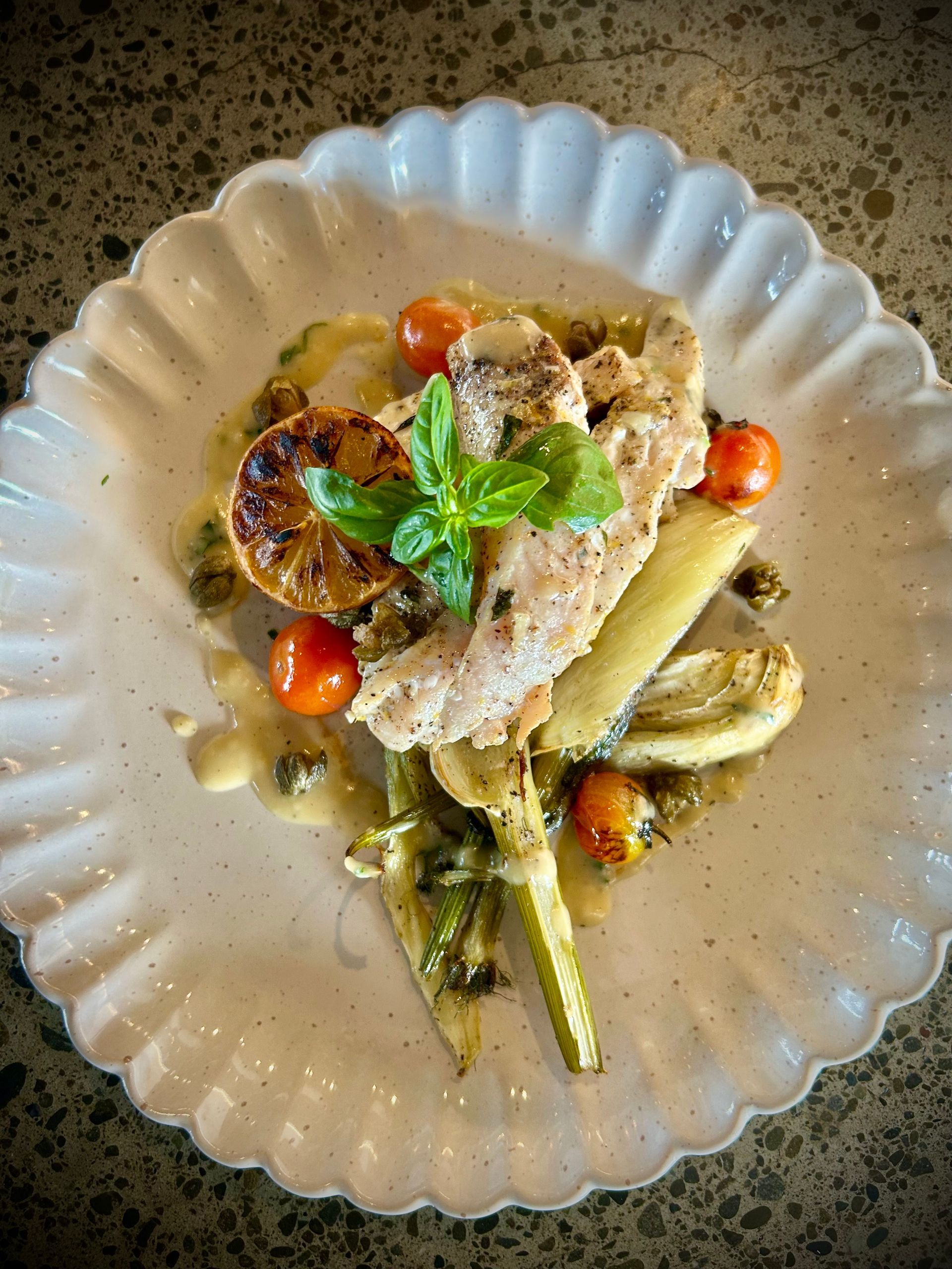 A grilled fish fillet topped with fresh basil, cherry tomatoes, and charred lemon on a white scalloped plate.