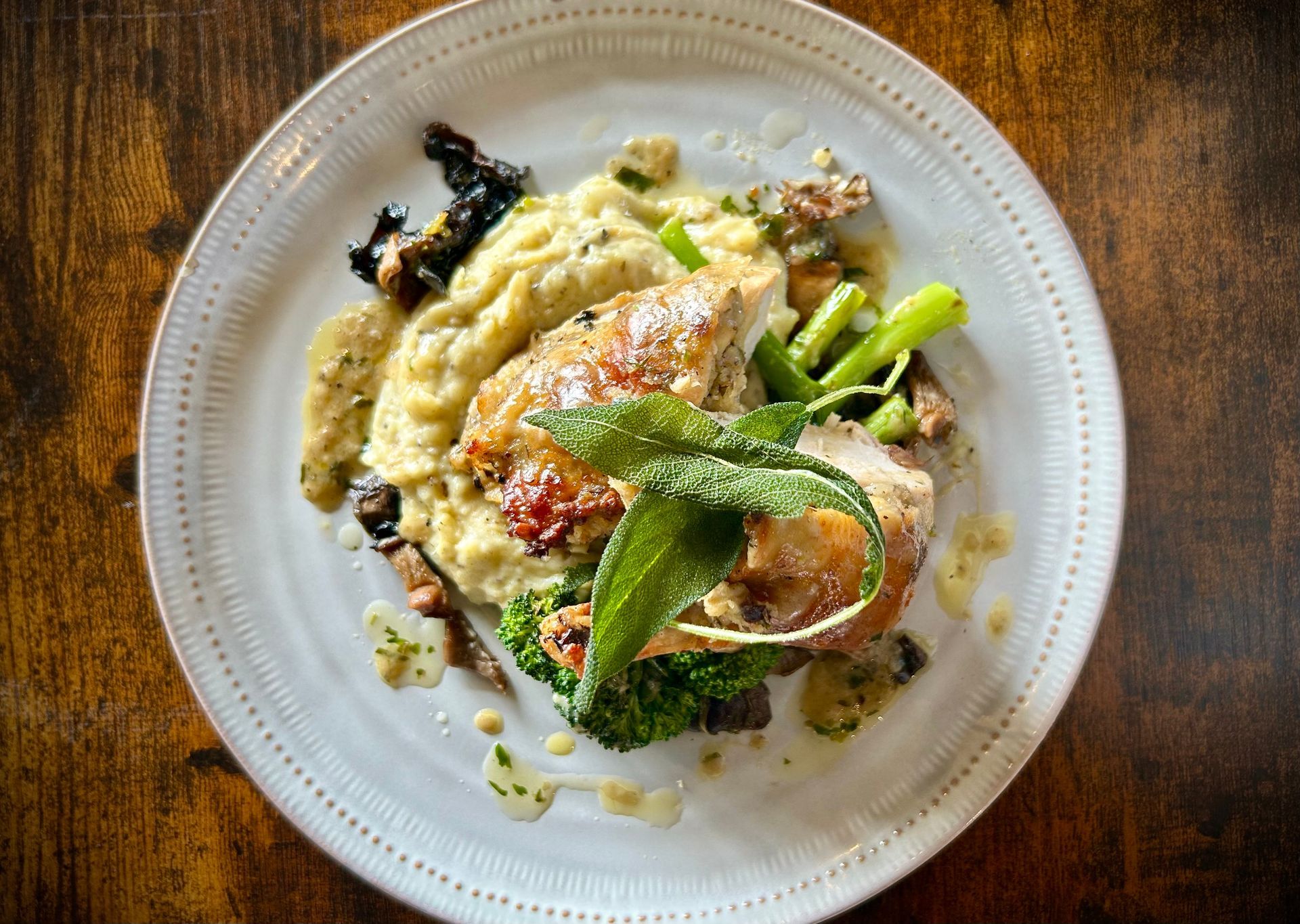 A top-down view of roasted chicken served with creamy polenta, charred broccoli, and crispy fried sage on a white plate.