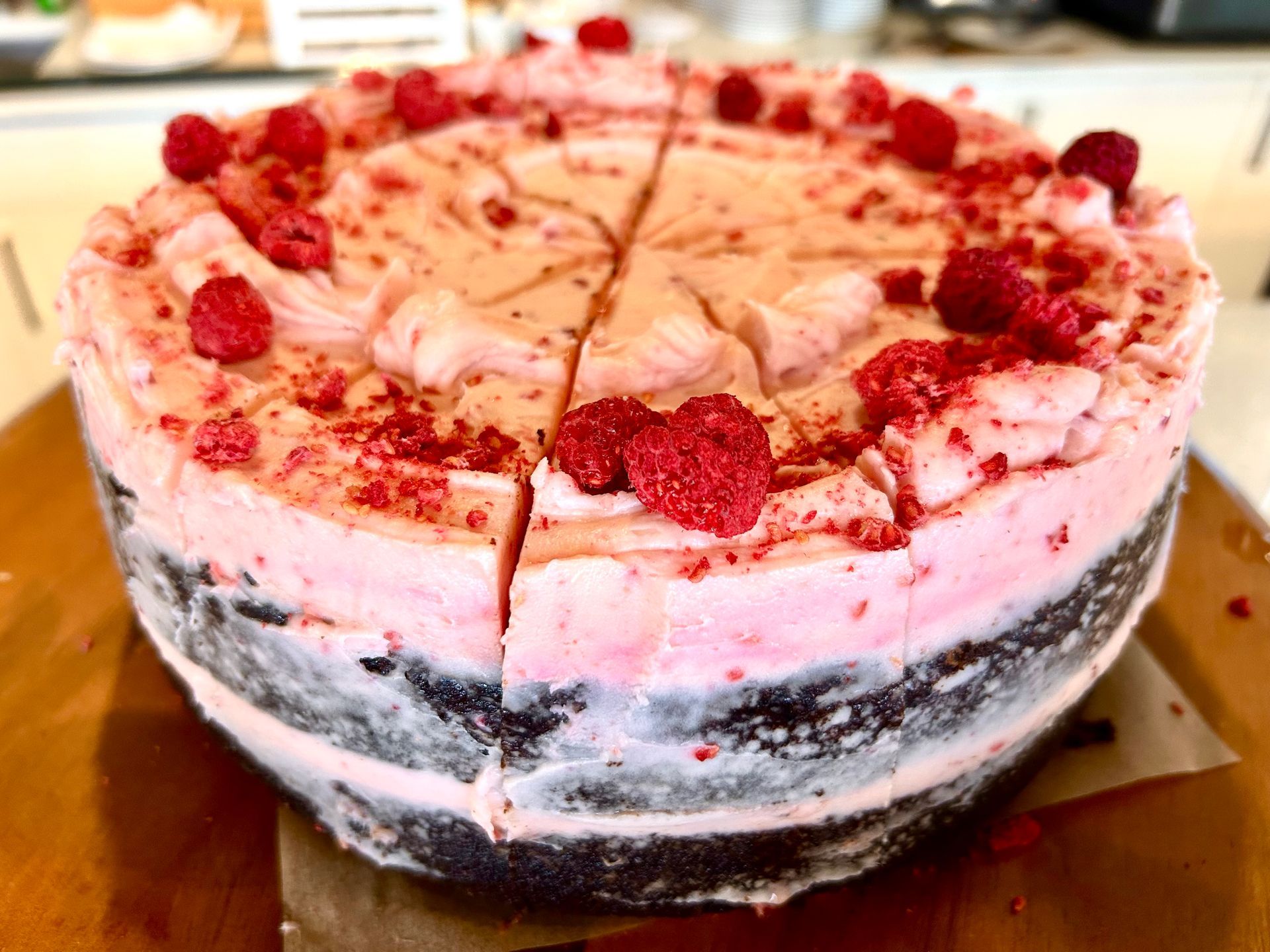 A layered chocolate cake with pink frosting, topped with fresh raspberries, sitting on a wooden board.