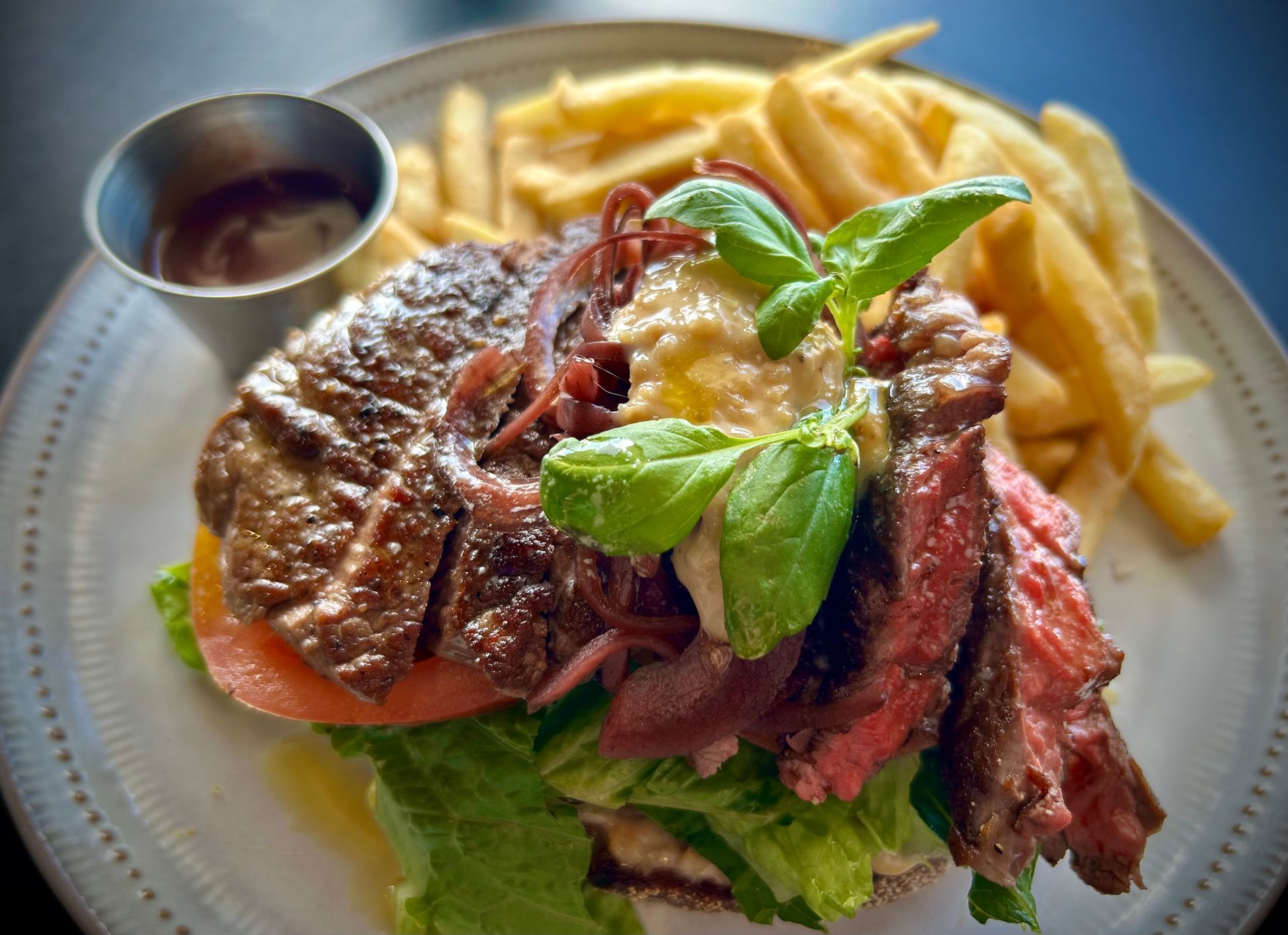 A steak sandwich with caramelized onions, greens, and fresh basil, served with french fries and a side of dipping sauce.