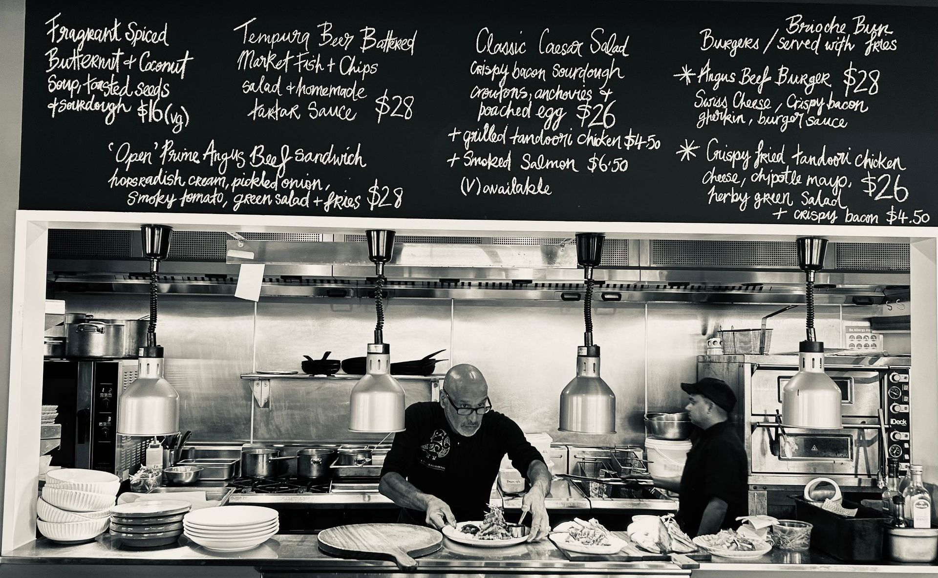 Two chefs work in a commercial kitchen under a blackboard menu, preparing food on a stainless steel counter.