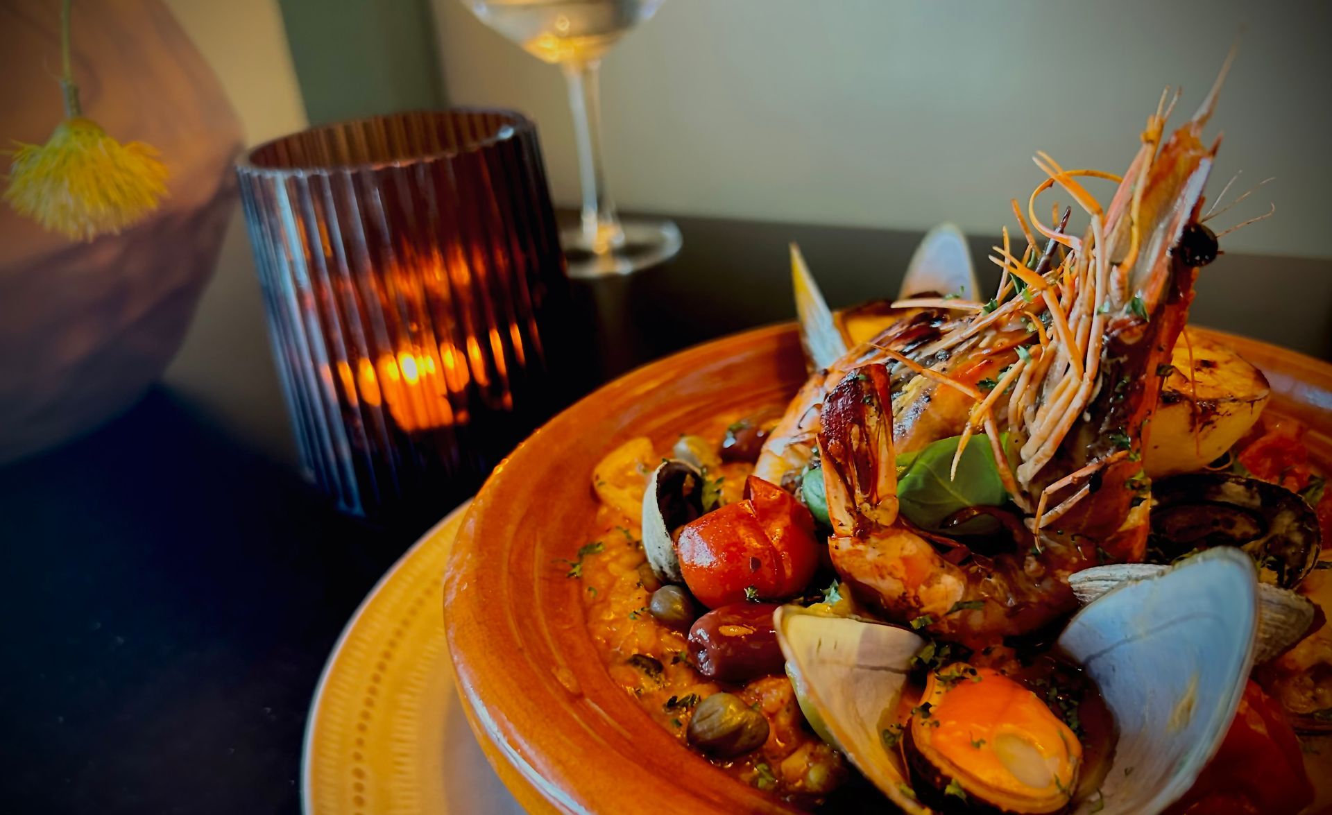 A close-up of a seafood dish with a large prawn and clams in an orange bowl, next to a lit candle on a dark table.