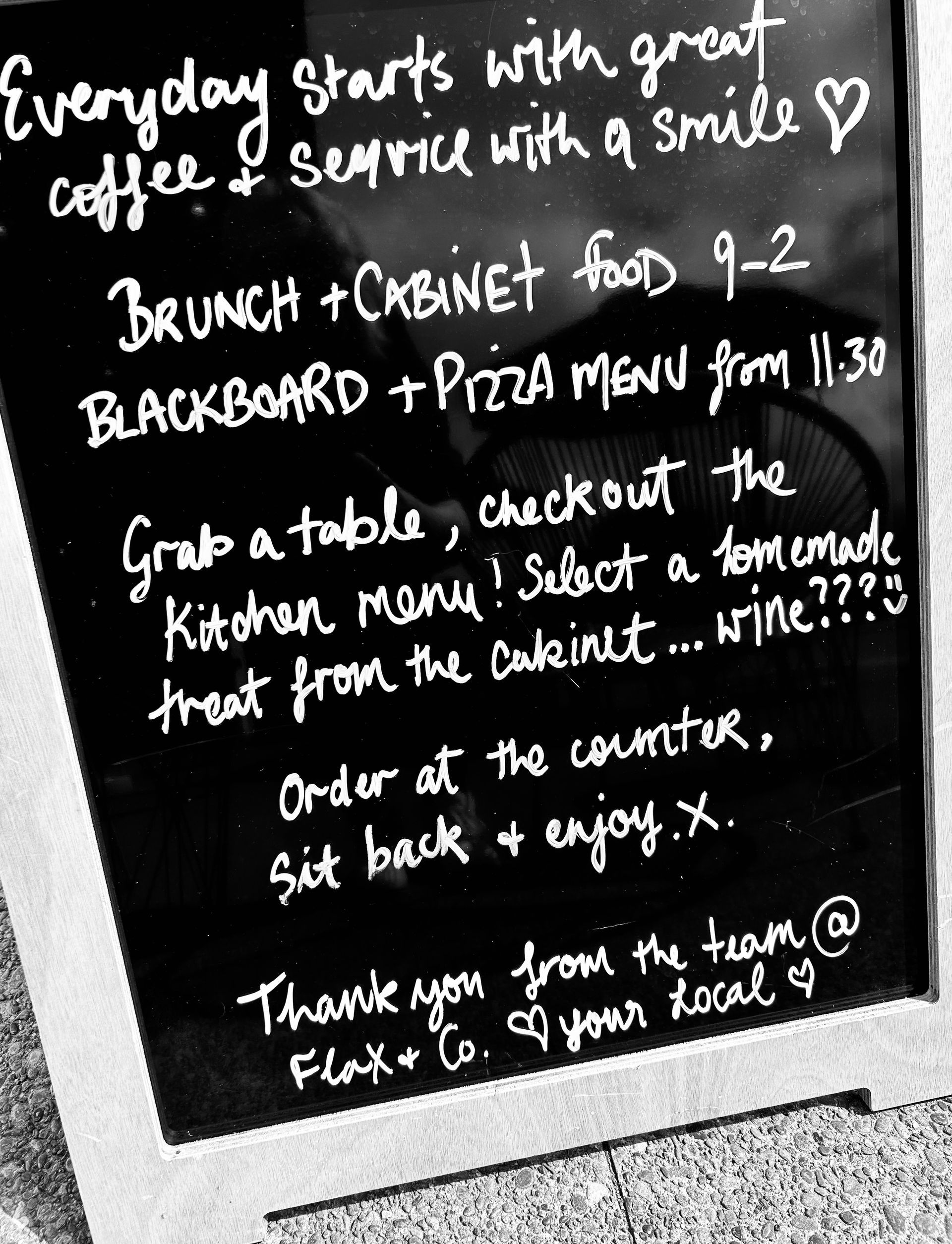 A chalkboard sign on an A-frame stand listing cafe menu hours, ordering instructions, and the name, Flax & Co.
