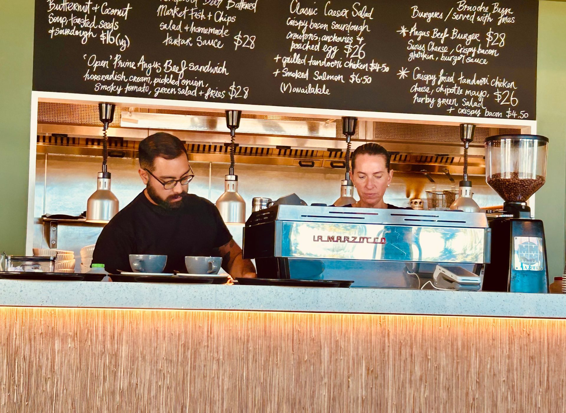 Two employees work behind a cafe counter with a chalkboard menu above them and a large espresso machine in front.