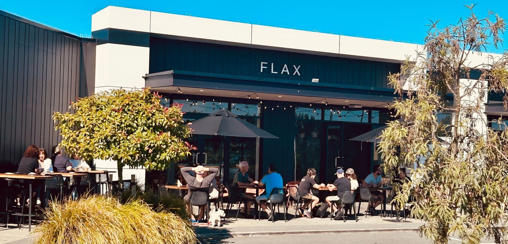 Patrons sit at tables under umbrellas on an outdoor patio in front of a modern storefront labeled 