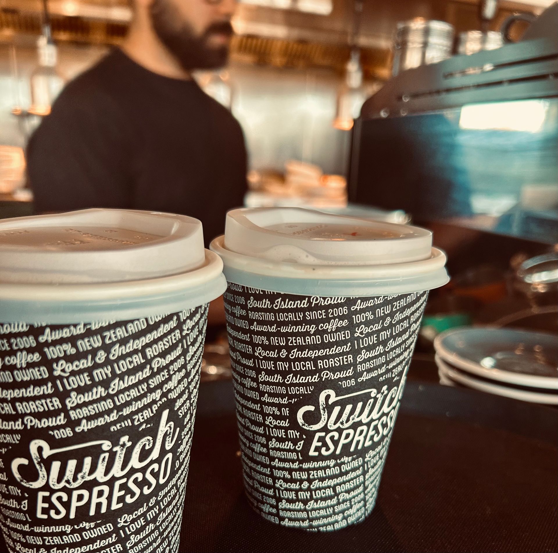 Two Switch Espresso coffee cups in the foreground, with a barista working behind a counter in a blurred coffee shop.