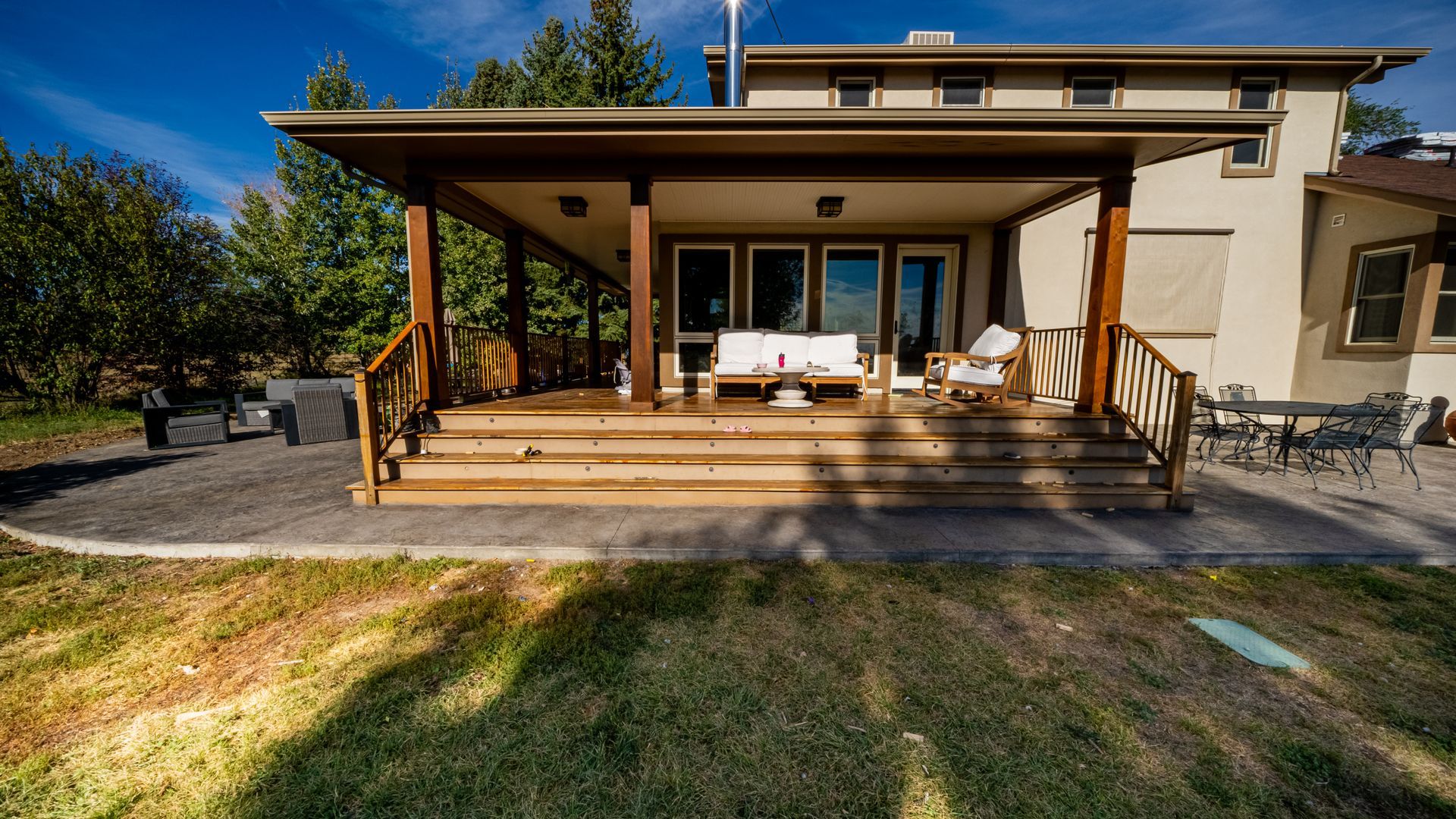 Covered wooden porch with steps, white seating, and glass doors on a modern house in a sunny yard