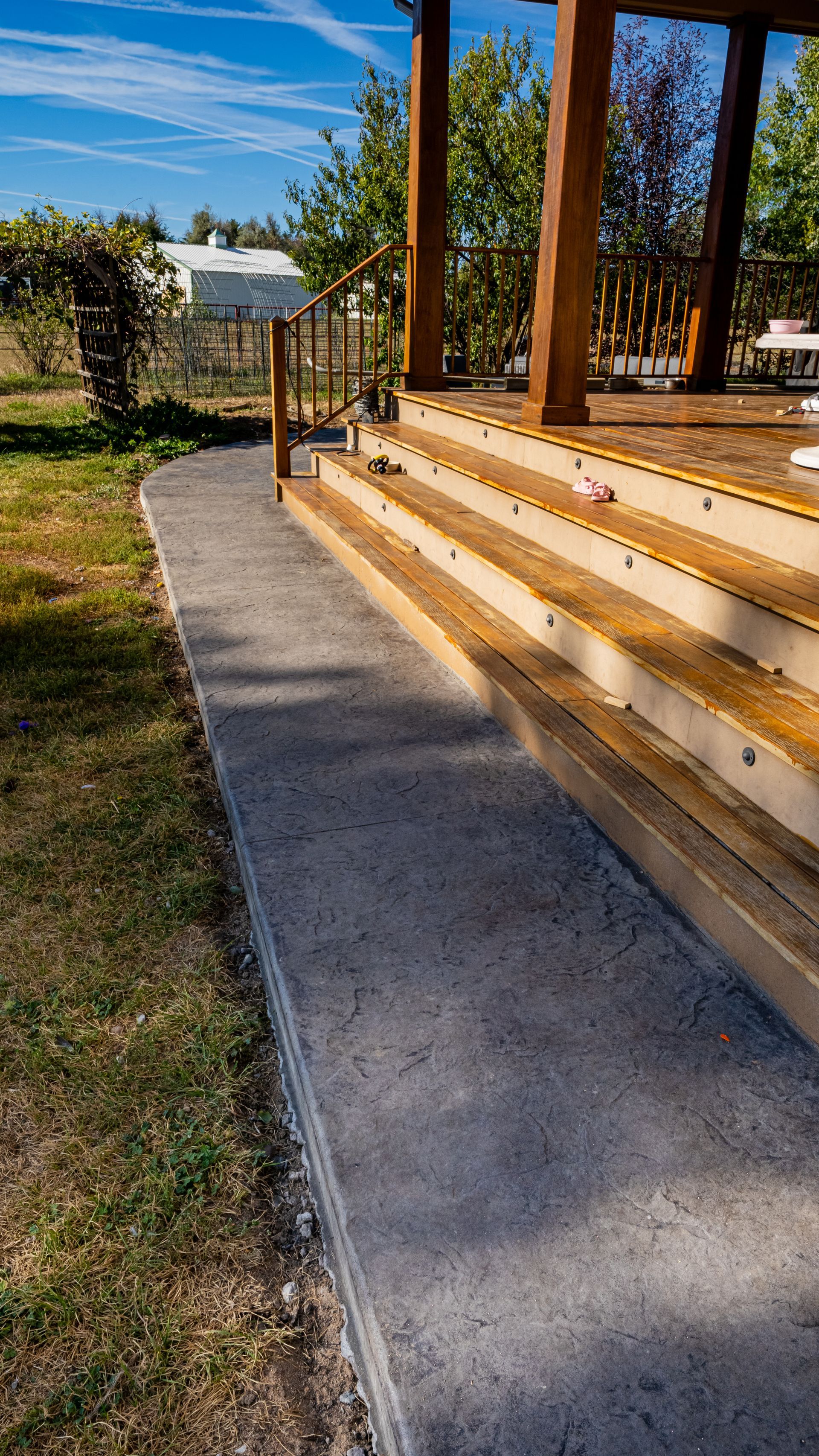 Concrete walkway beside wooden deck steps in a sunny yard with trees and grass