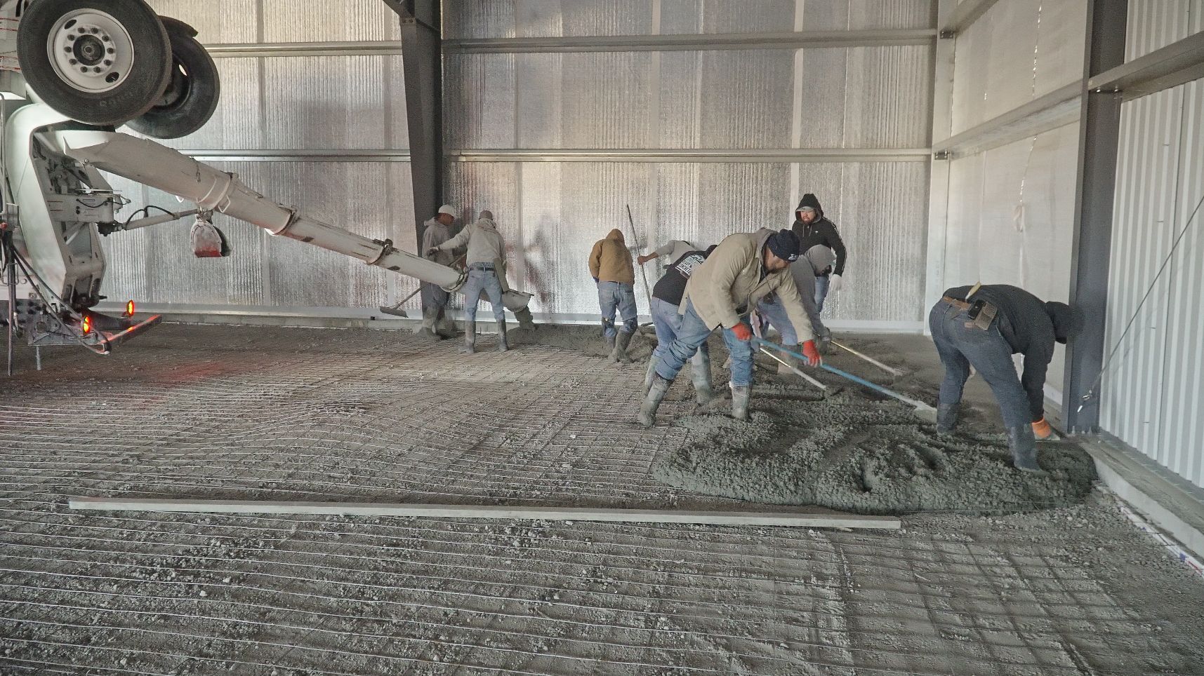 Workers smoothing wet concrete inside a large metal-walled construction site, with a mixer truck at left