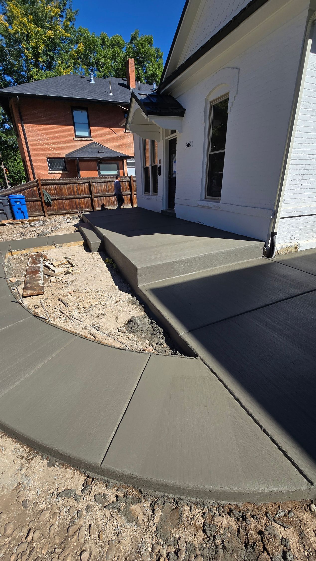 Curved concrete walkway beside a white house under construction, with dirt and a brick building in the background.
