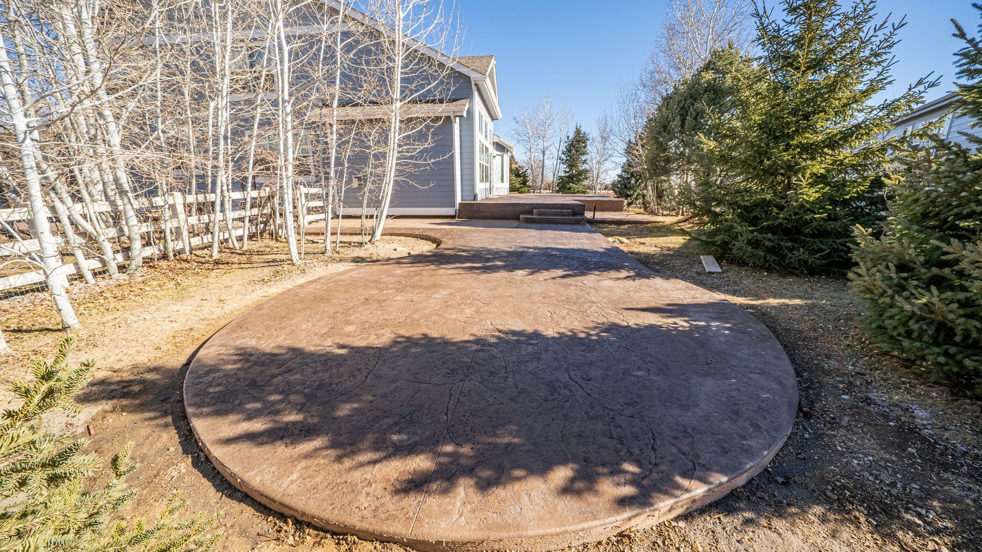 Round concrete patio in a yard beside a house, surrounded by trees and shrubs on a sunny day