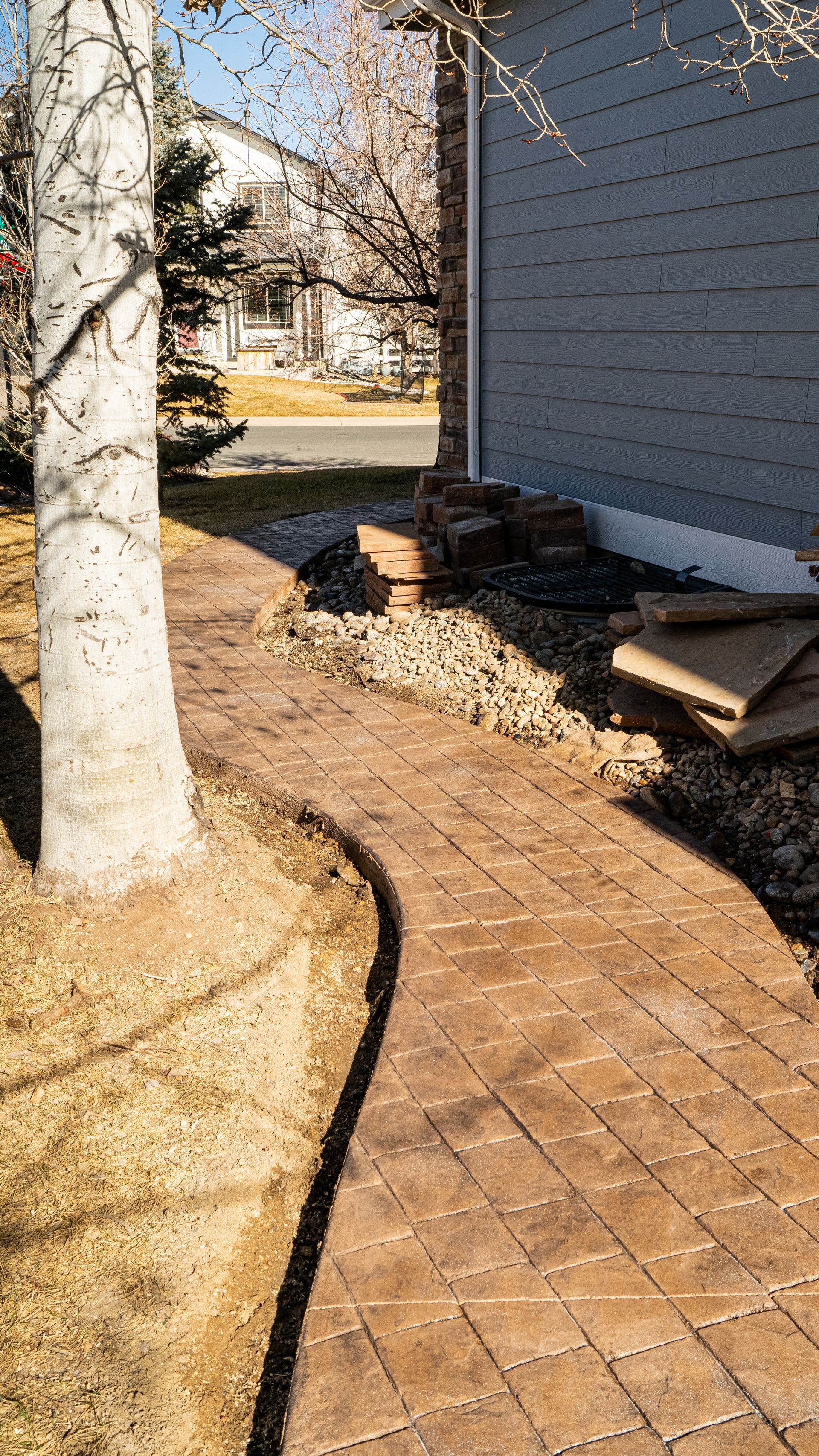 Curved brick walkway beside a house, with a tree, mulch bed, and stacked boards along the wall.