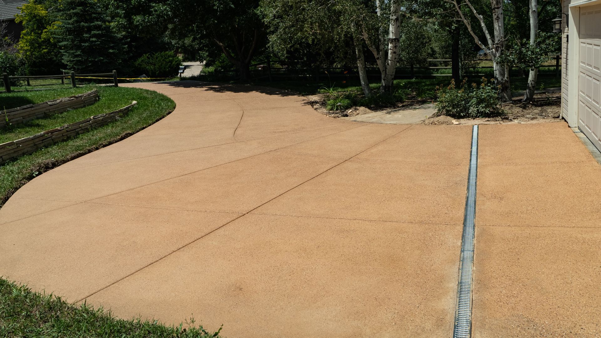 Curved tan concrete driveway beside a house, bordered by grass and trees