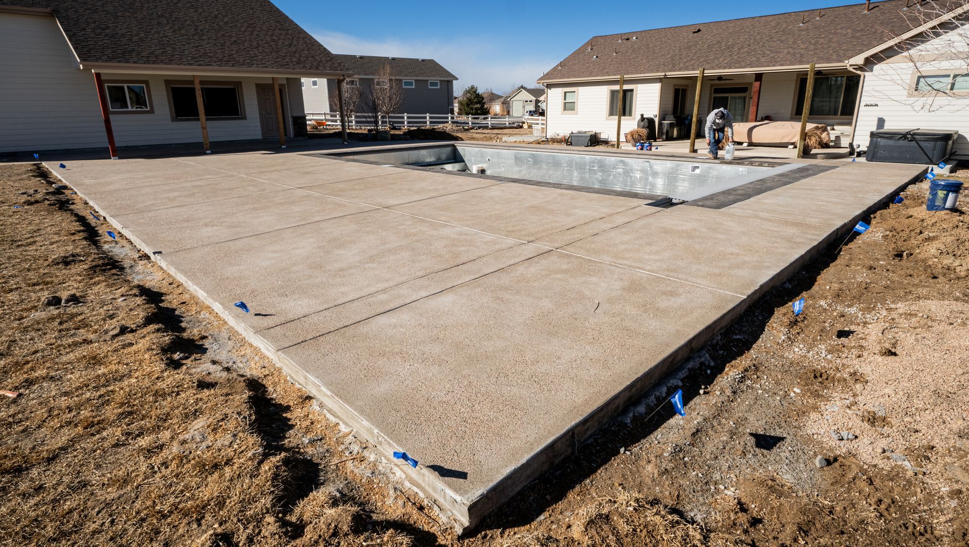 Fresh concrete patio in a backyard between two houses, bordered by dirt and blue stakes.