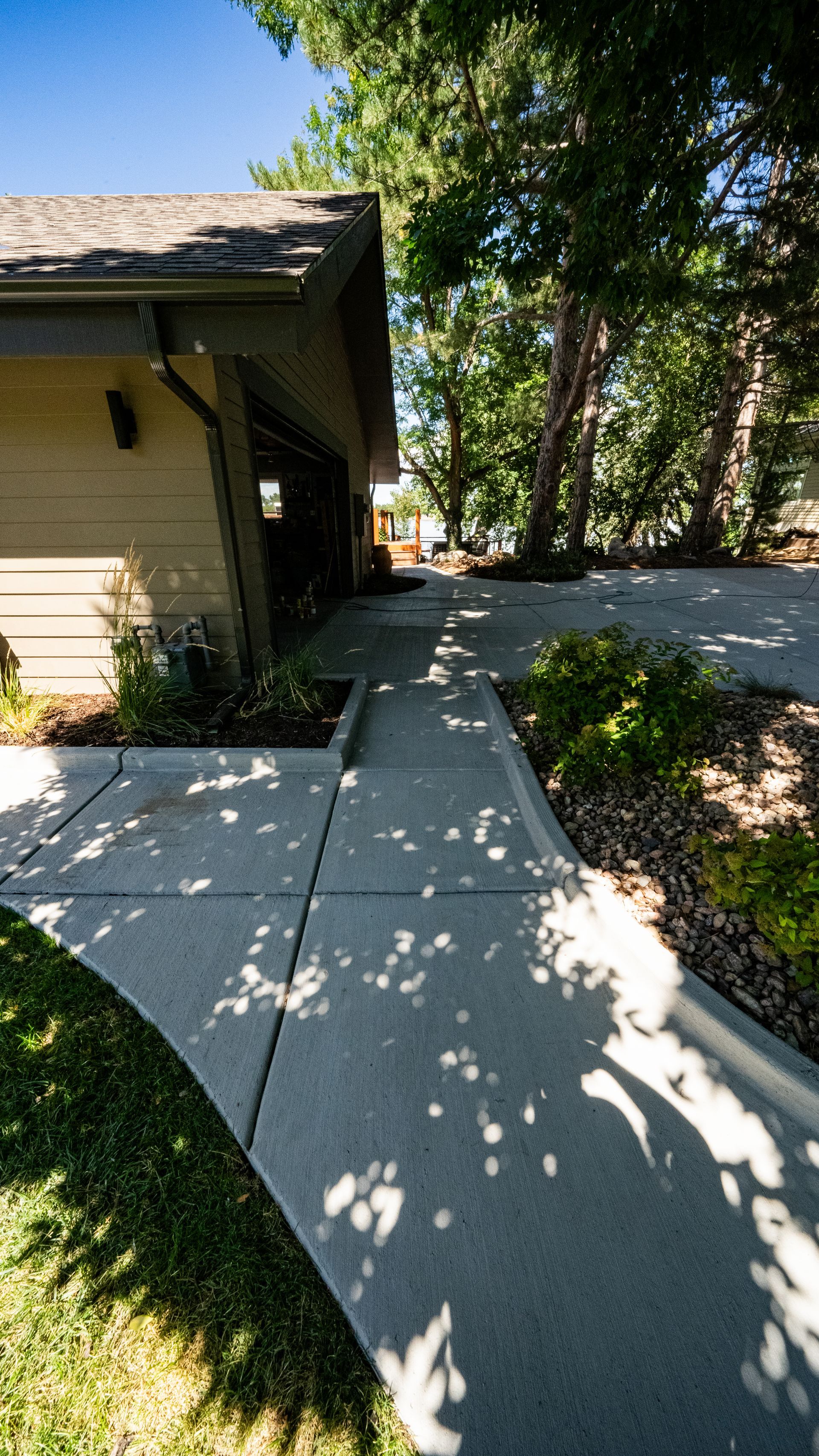 Concrete walkway beside a house, with trees casting dappled shade and a driveway in the background