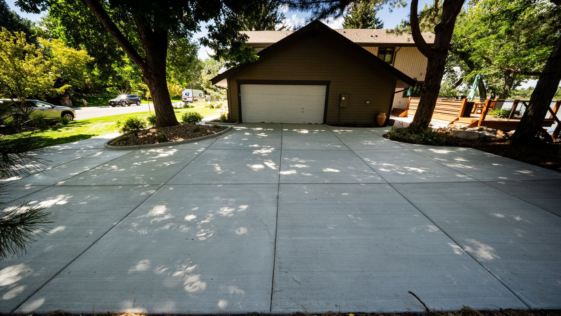 Wide concrete driveway leading to a brown garage under trees in a sunny suburban yard