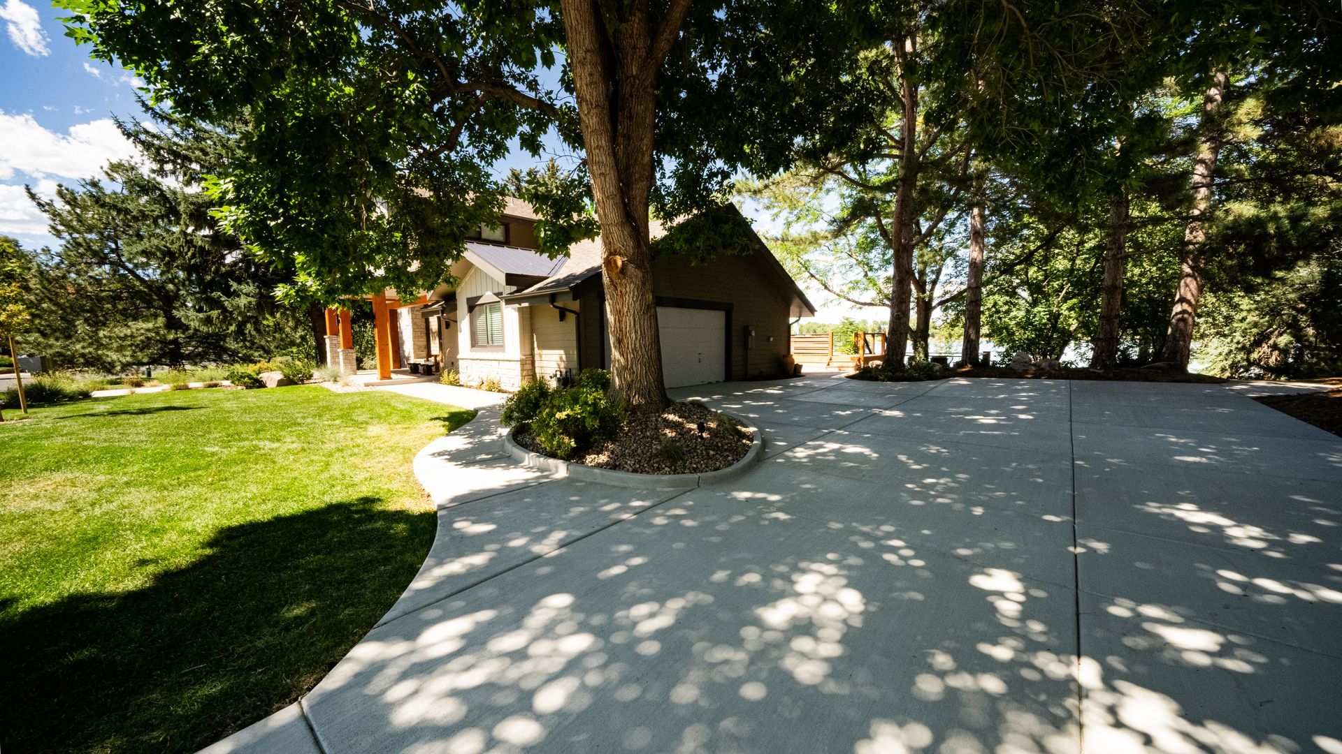 Stone driveway beside a house under trees, with a grassy lawn and bright sunlit yard.