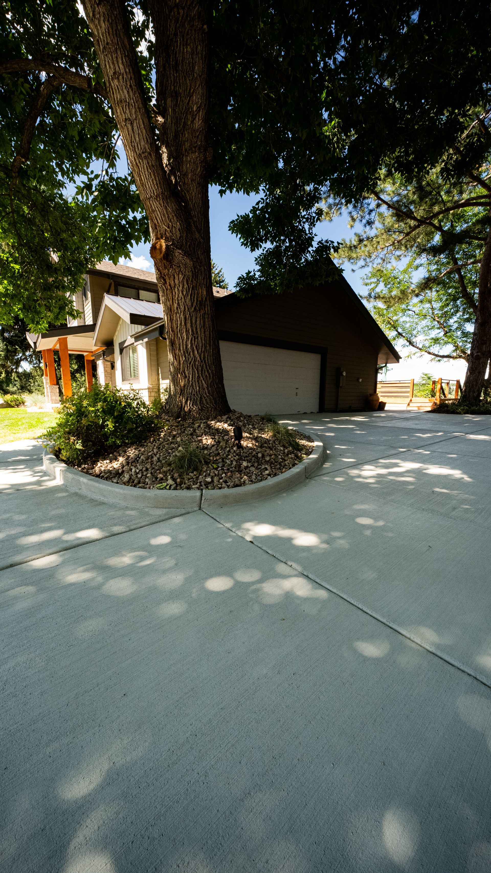 Tree-shaded driveway beside a small house and garage, with gravel and sunlight filtering through leaves