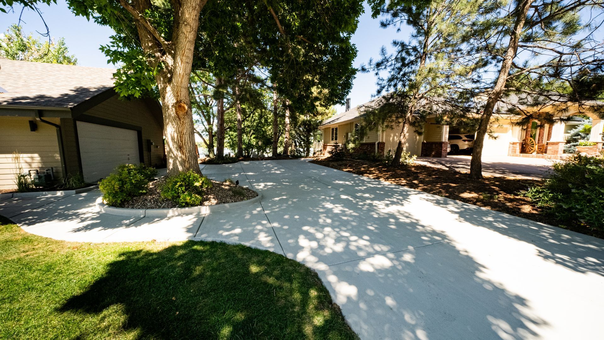 Sunny residential driveway shaded by trees, leading past houses with a manicured lawn.