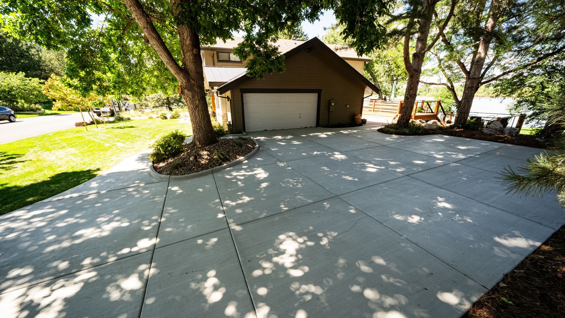 Wide concrete driveway leading to a garage under trees, with dappled sunlight and landscaped yard