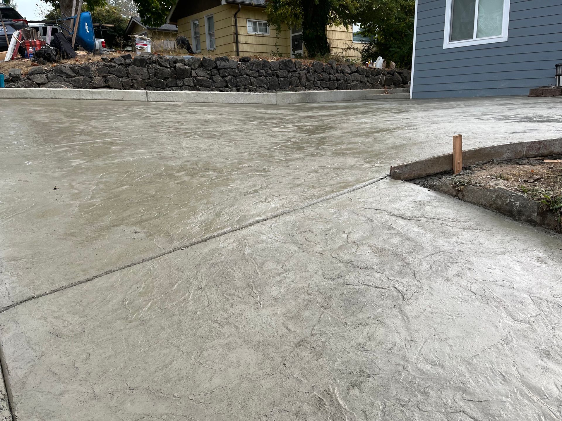 Concrete driveway leading to a house, with a stone retaining wall and yard in the background