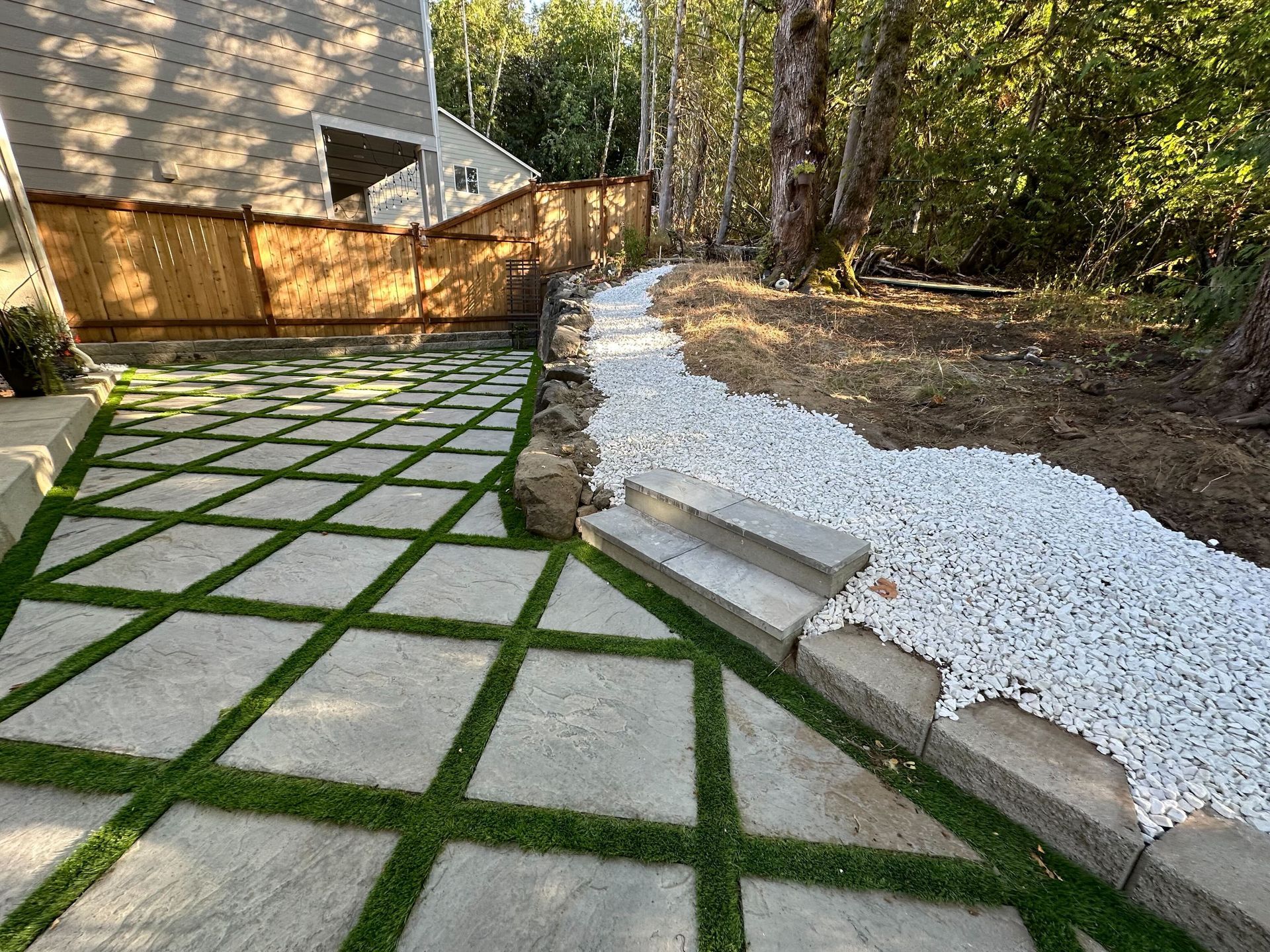 Modern backyard patio with geometric pavers, white gravel, stone edging, and a wooden fence near trees