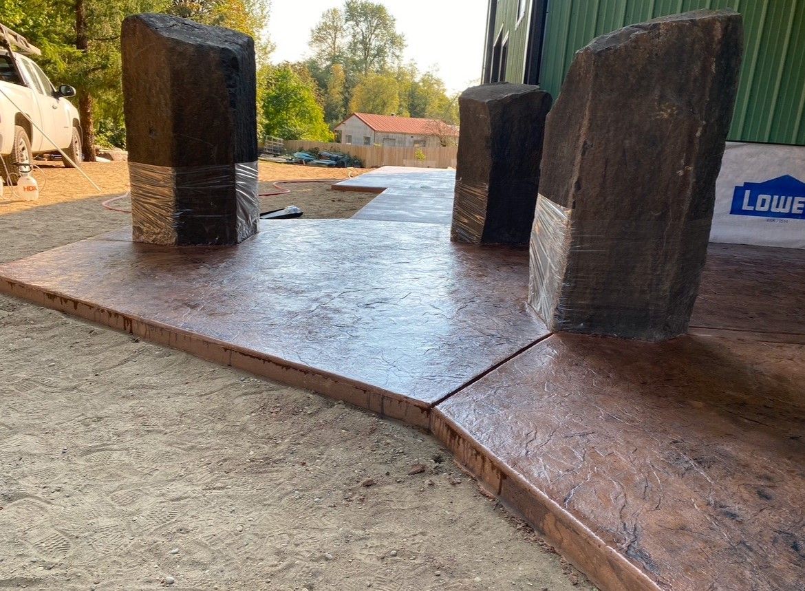 Rust-colored concrete walkway with three dark stone pillars in a landscaped outdoor area