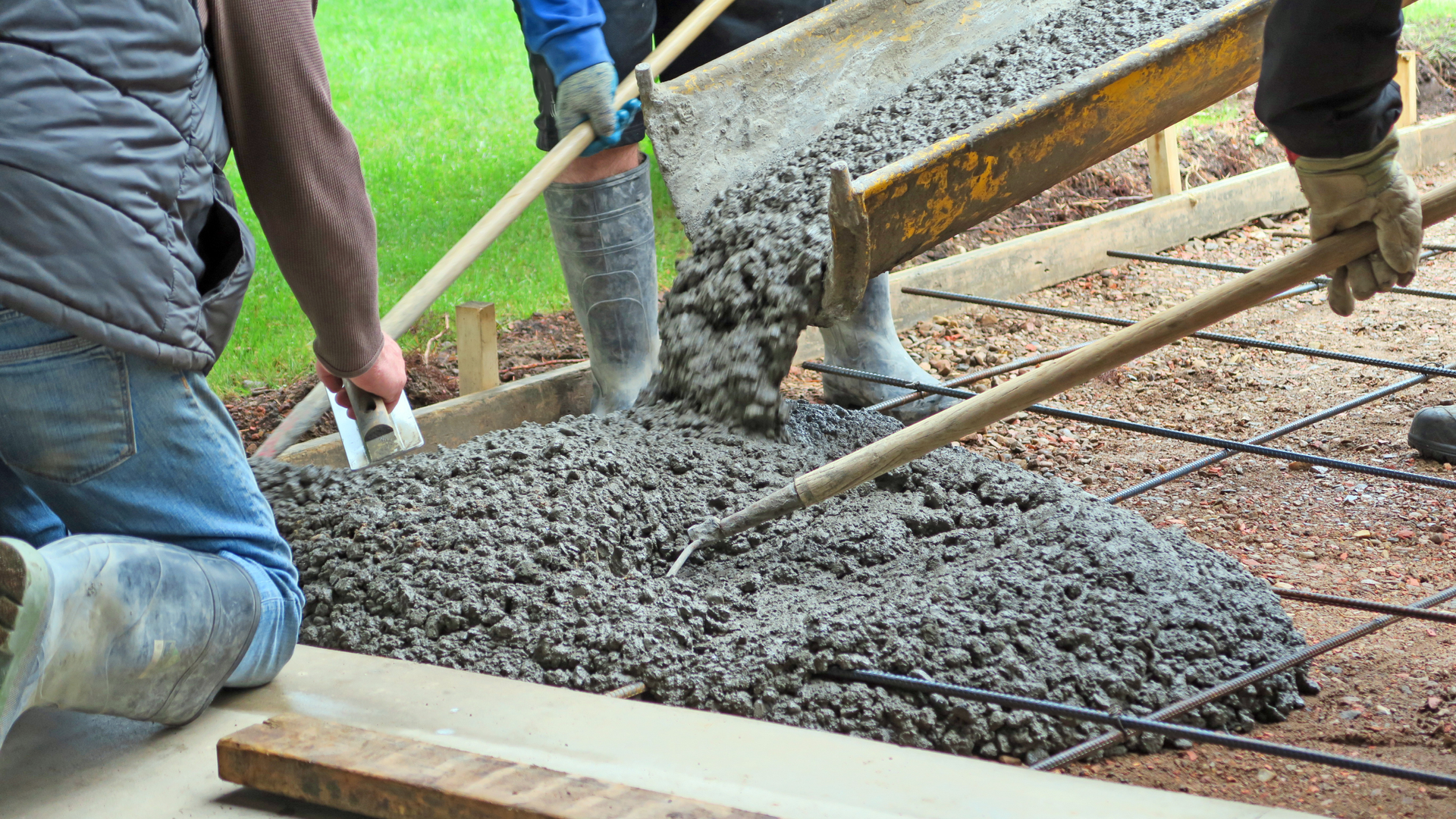 Workers pouring wet concrete with shovels and a hose over a rebar grid at a construction site
