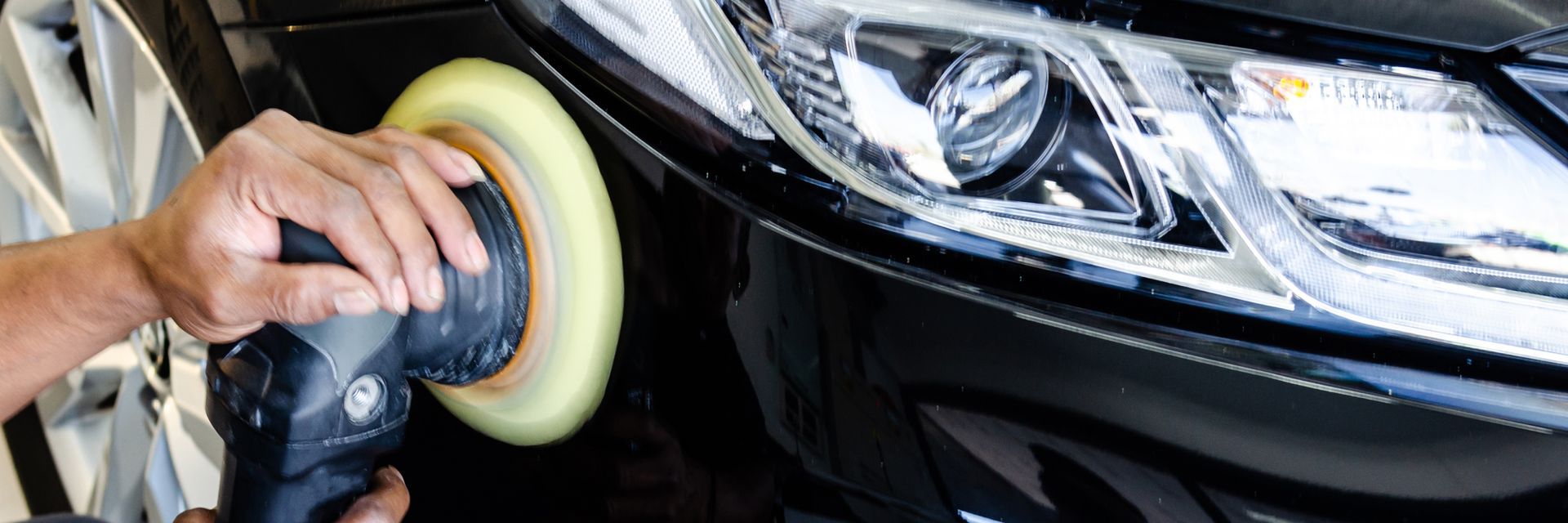 Person polishing a black car headlight with a buffer, close-up of the front bumper