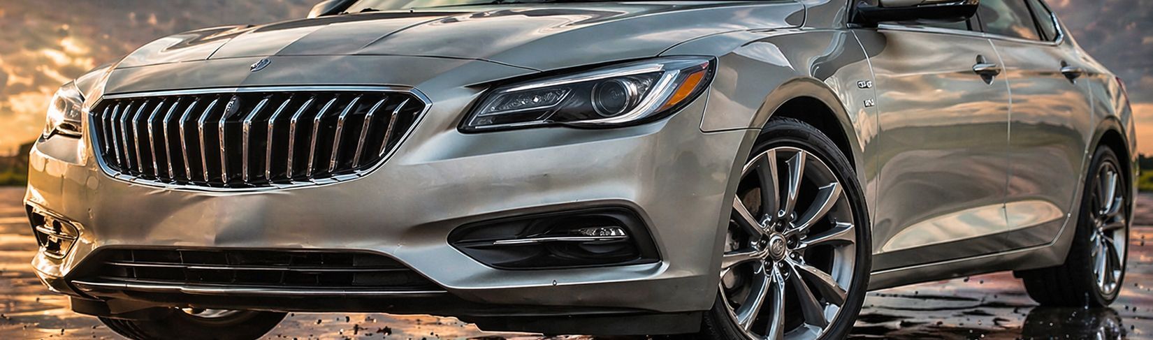 Silver luxury sedan parked on a wet street, front view with chrome grille and headlights lit.