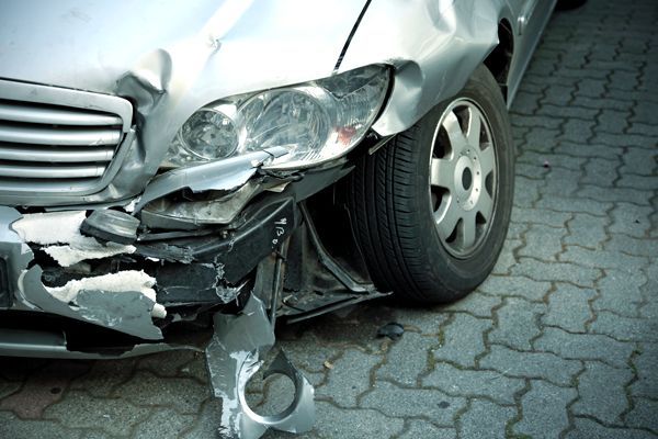 A silver car with a broken bumper is parked on a brick driveway.