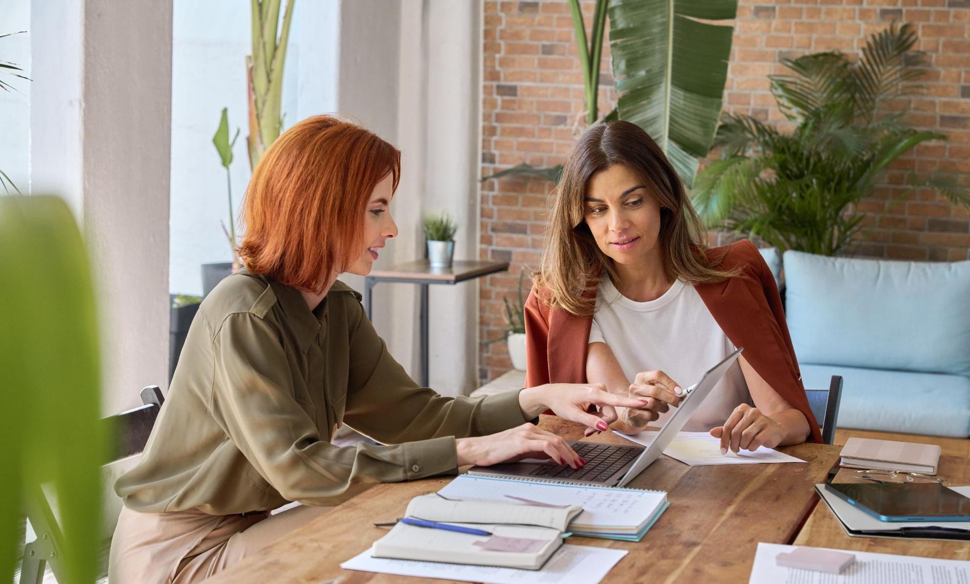 Two women reviewing laptop at wooden table in a bright office.