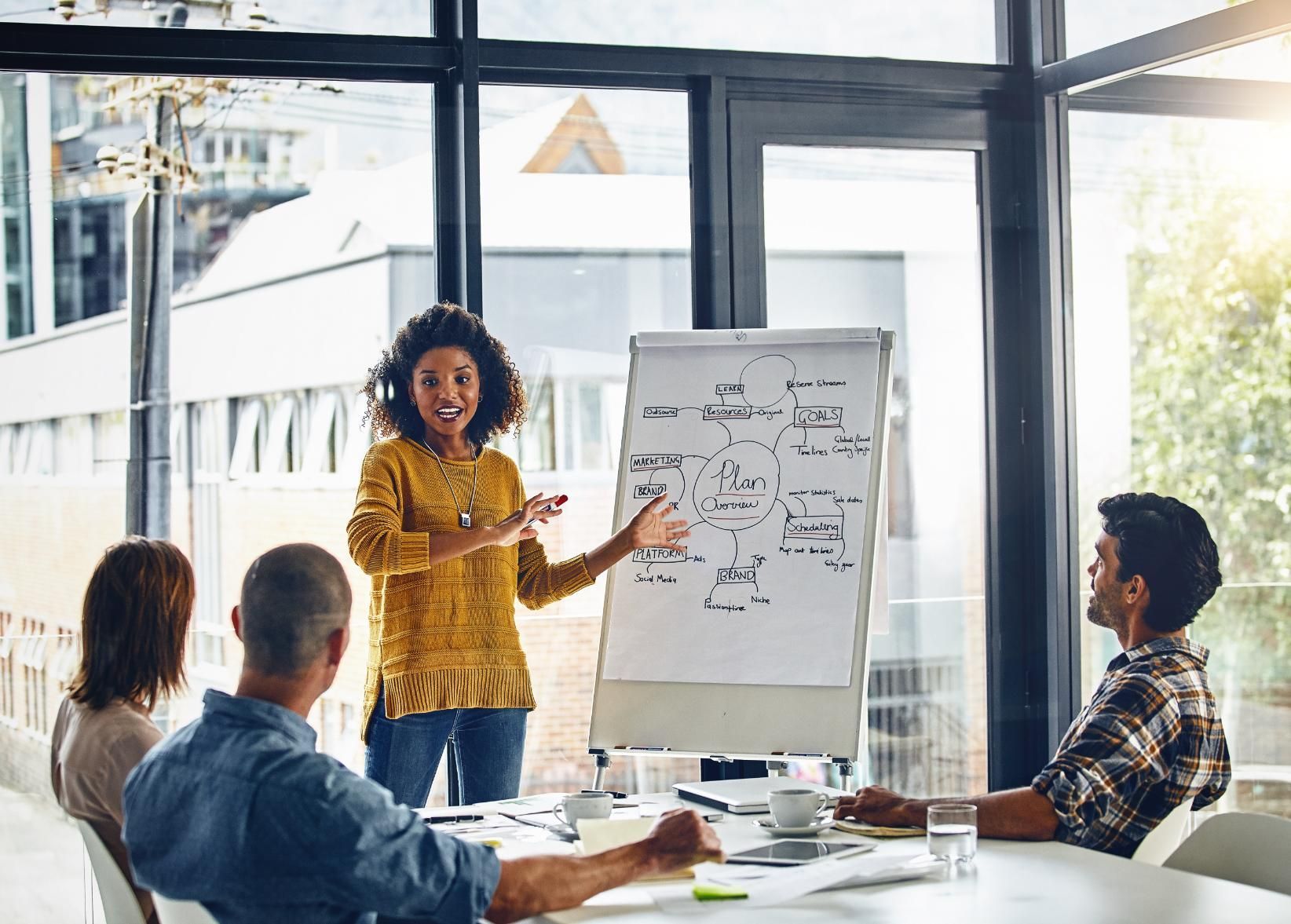 A woman presents a whiteboard to a team in a sunlit office, gesturing and smiling. 