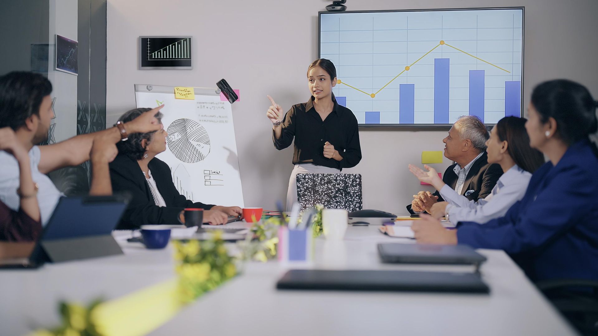 A woman leads a presentation in a conference room, gesturing to a whiteboard while colleagues observe and discuss, with graphs displayed on the wall.
