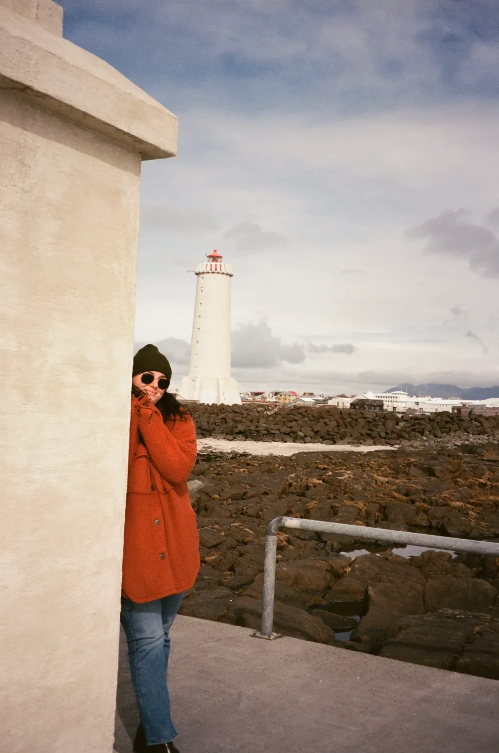 Film photograph of a woman in orange coat beside a white structure, lighthouse in background, cloudy sky.