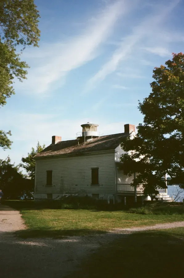 Old white house with a cupola, set on a grassy hill under a bright blue sky. Trees frame the sides.