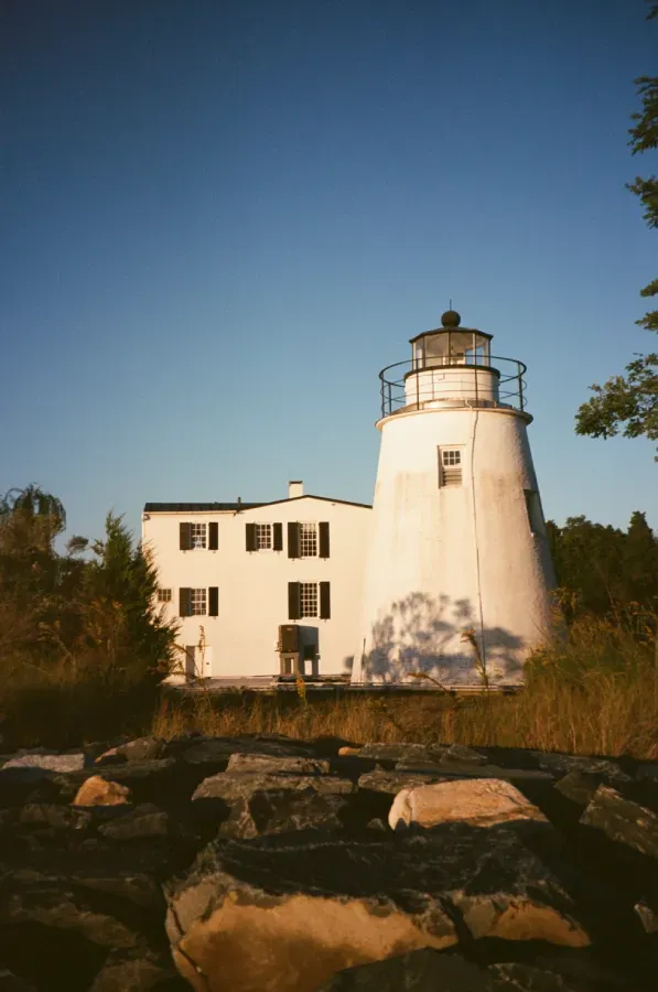 White lighthouse with attached building against a clear blue sky. Rocky foreground, trees in the background.