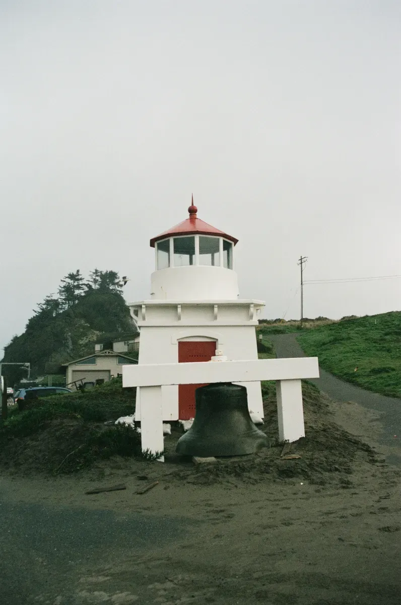 A small white lighthouse with a red roof stands outdoors next to a large, dark bell hanging from a white wooden frame.