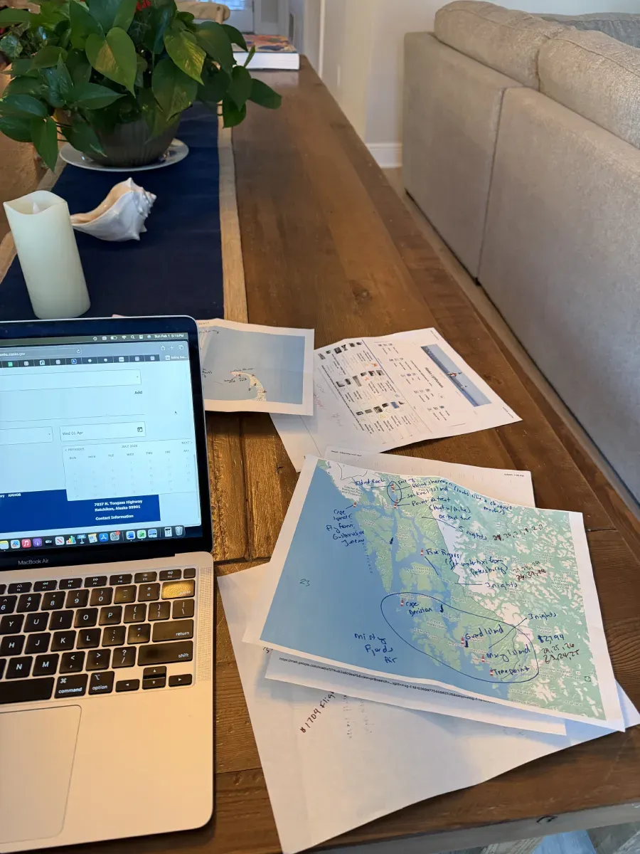 A laptop sits on a wooden table alongside printed papers, charts, and a plant, suggesting a workspace or study setup.