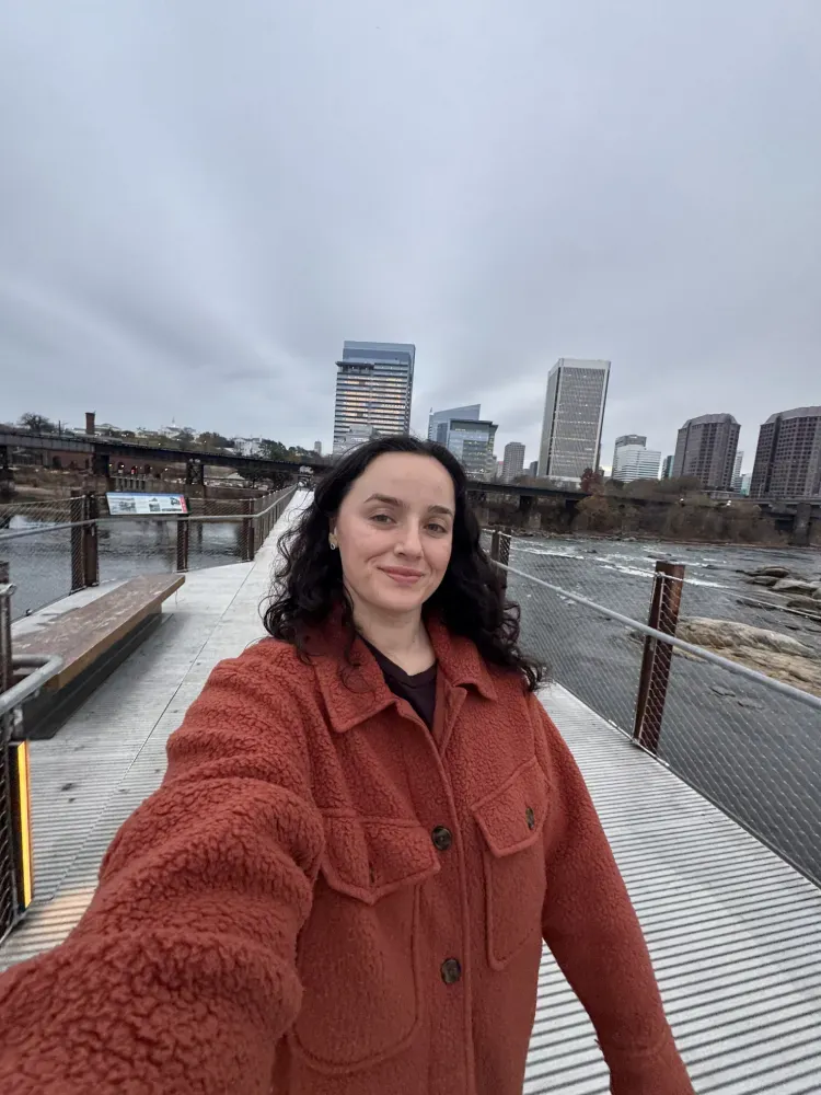 Woman taking a selfie on a dock by the water, wearing a rust-colored coat with a city skyline behind her.