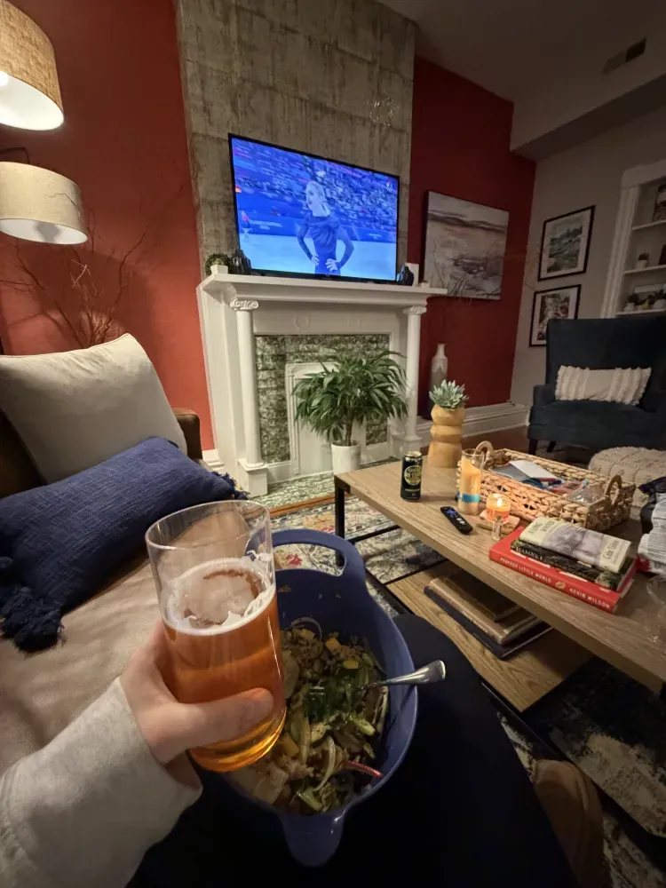 Person holding a beer and salad in a cozy living room with TV and coffee table snacks
